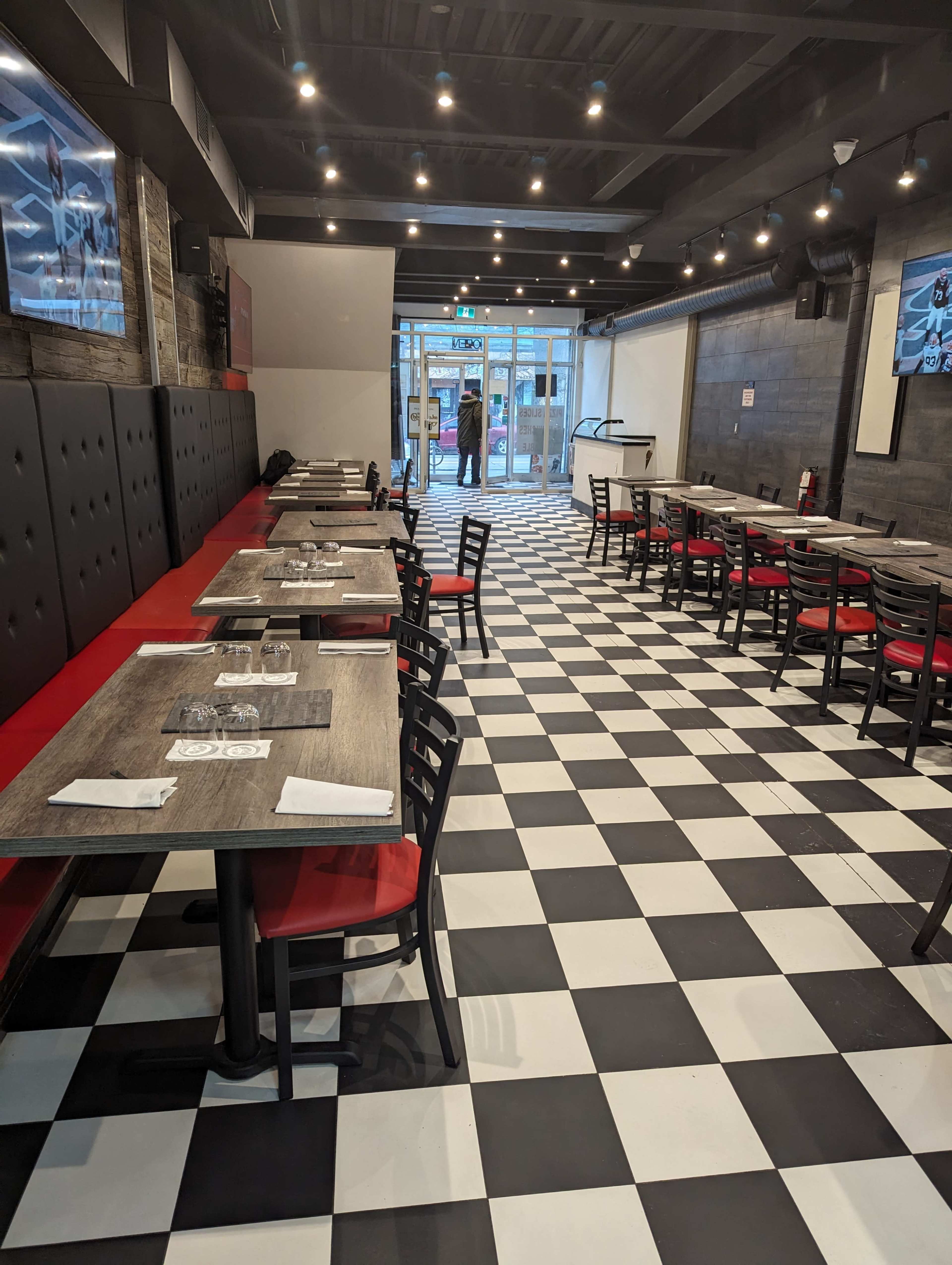 A restaurant interior with a black and white checkered floor, red upholstered seating, and multiple empty tables arranged in rows.