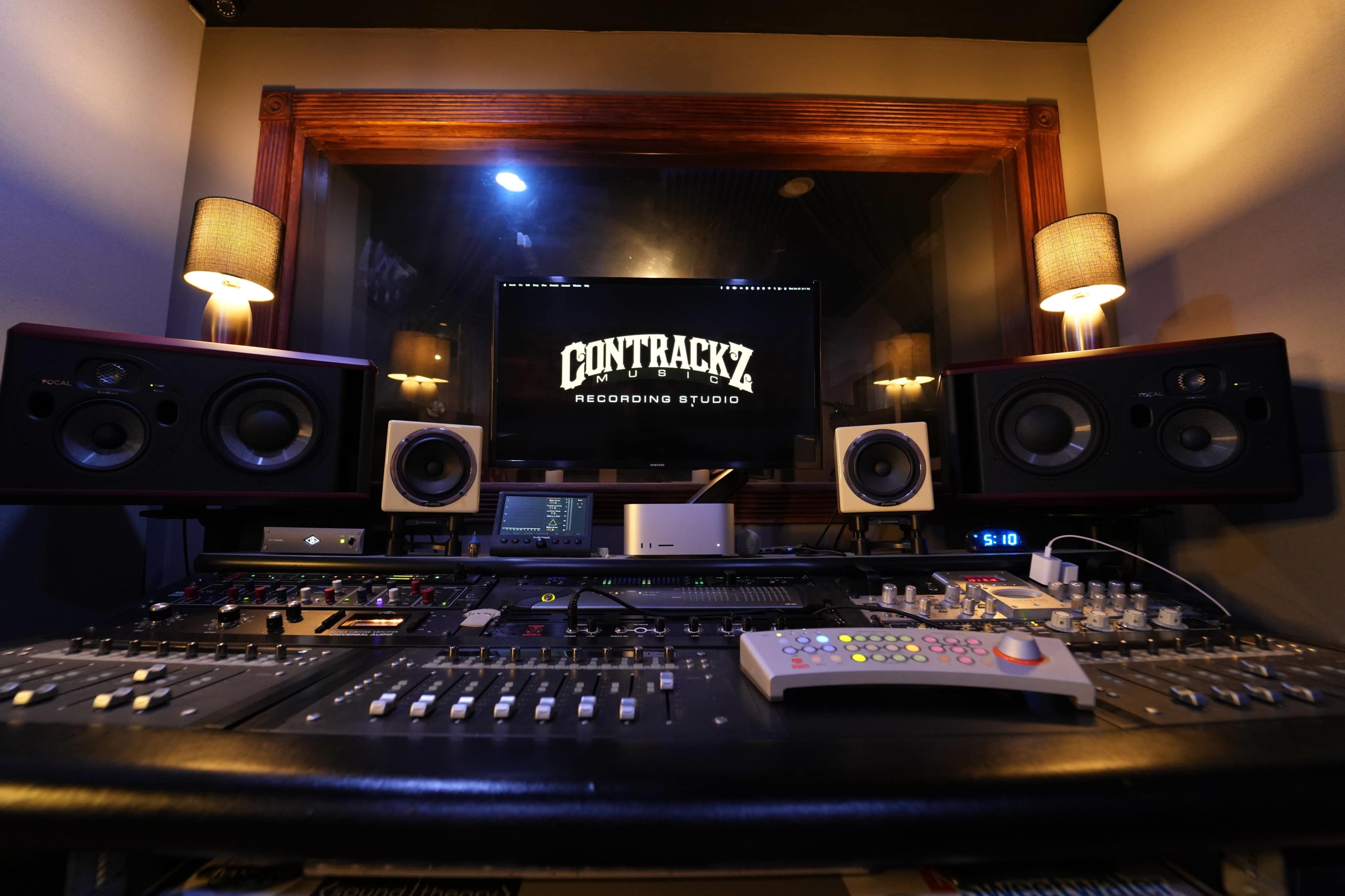 The image shows a recording studio with a digital audio workstation, studio monitors, and a control surface, illuminated by soft lighting.