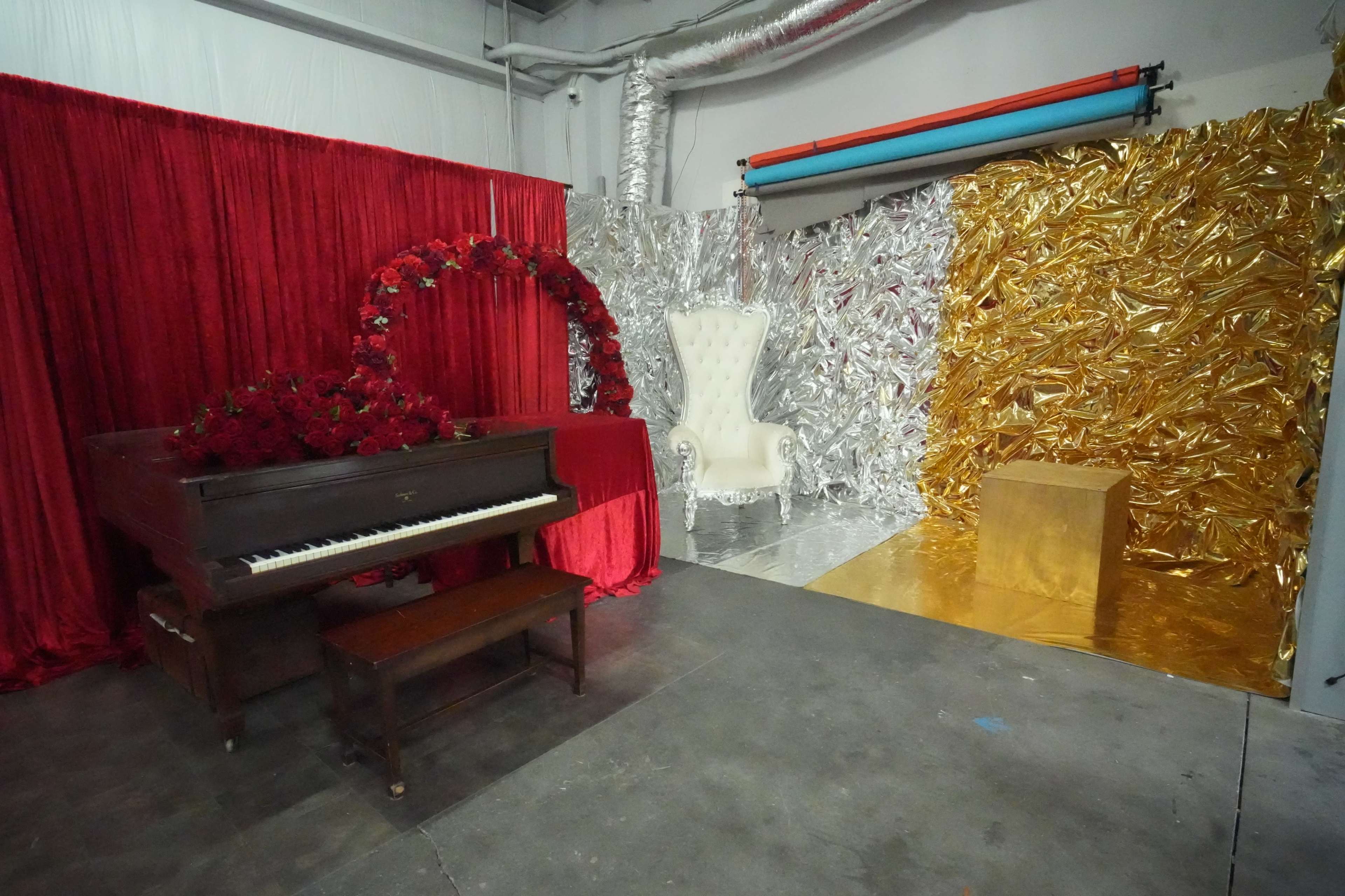 The scene features a vintage piano with a small wooden bench, surrounded by red curtains and floral decorations, next to a shiny gold and silver backdrop with a white, ornate chair nearby.