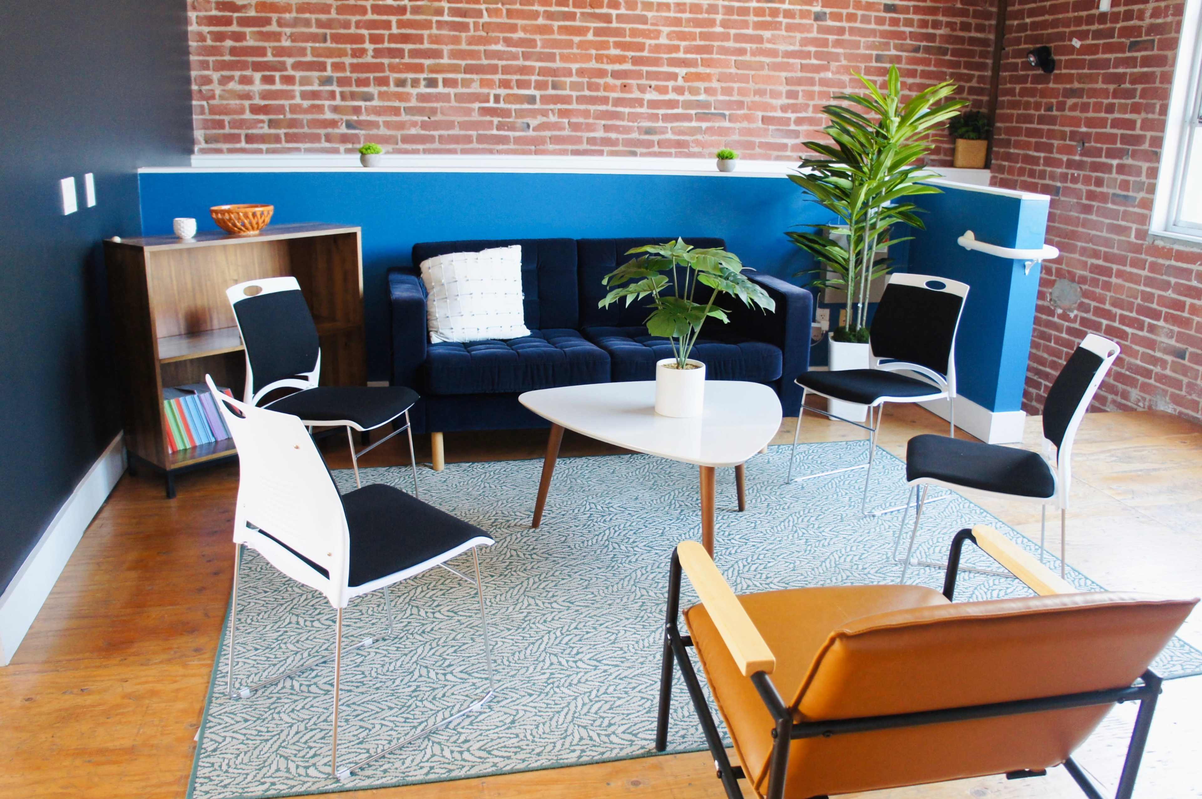 The image shows a modern lounge area with a blue and brick wall, featuring a dark blue sofa, a white coffee table, several chairs, and a potted plant.