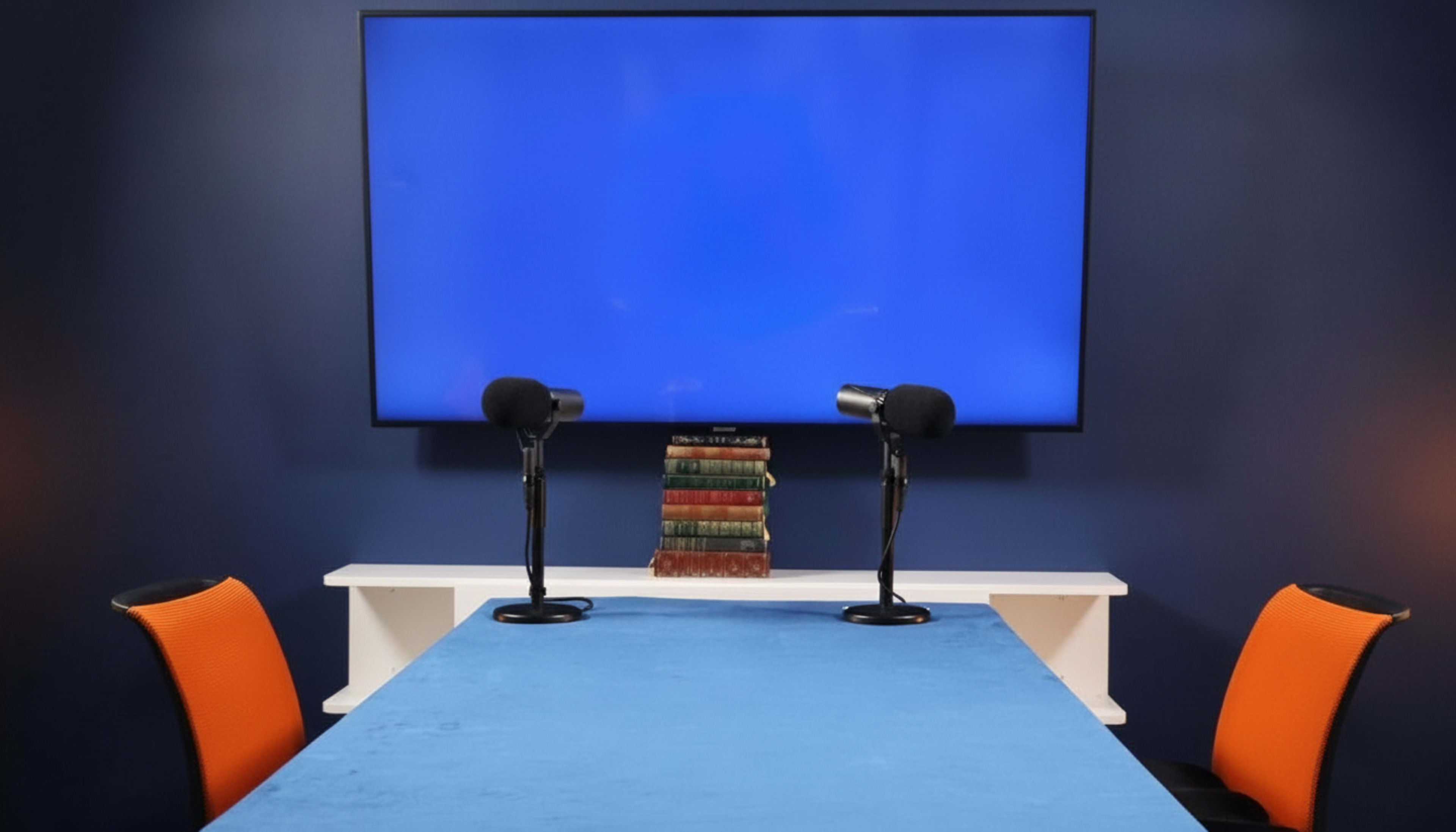 A blue wall features a large television screen, while a table with a blue cover is flanked by two chairs and topped with a stack of books and two microphones.
