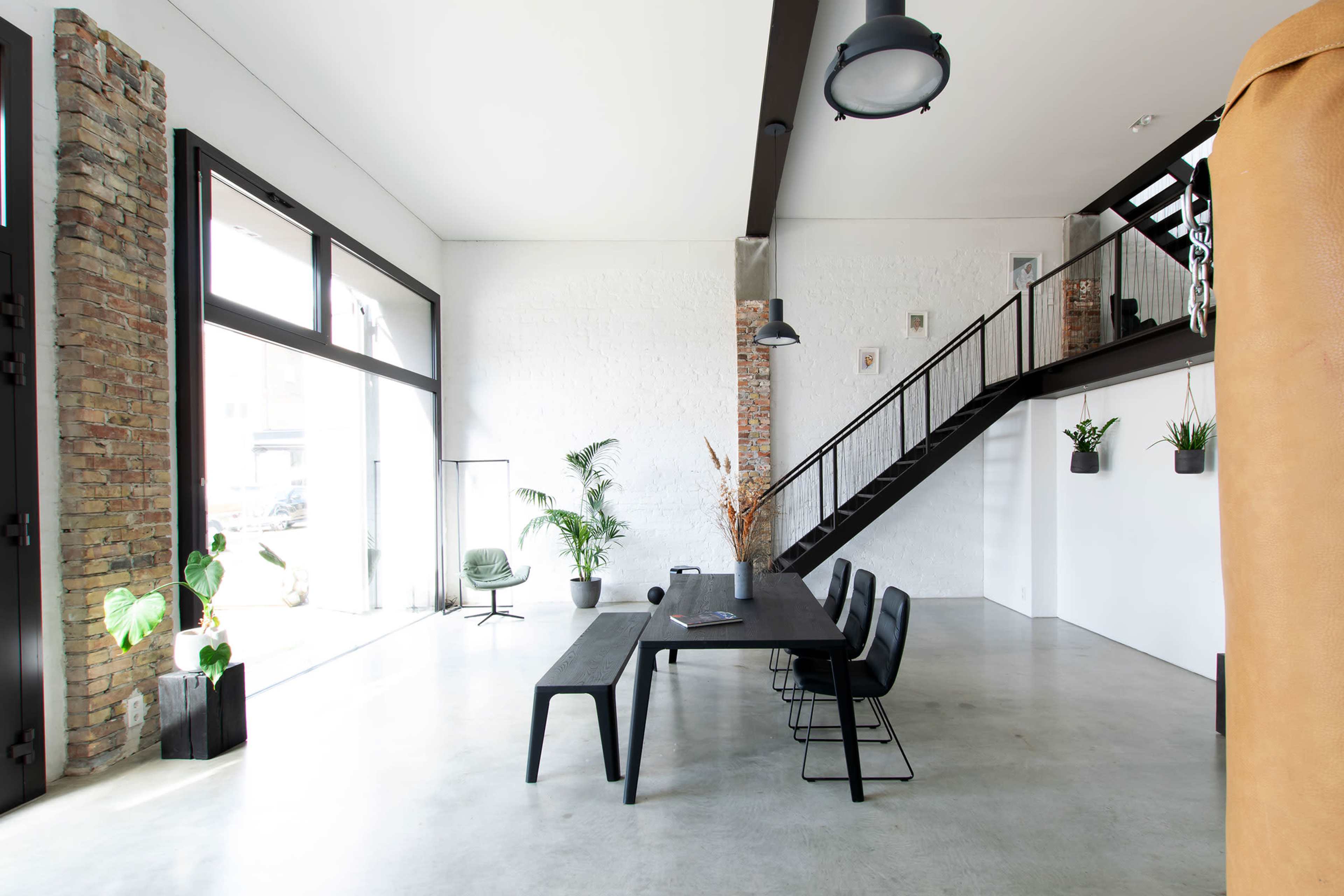 A modern interior space featuring a large dining table with six chairs, a staircase, and several plants against a backdrop of exposed brick and white walls.