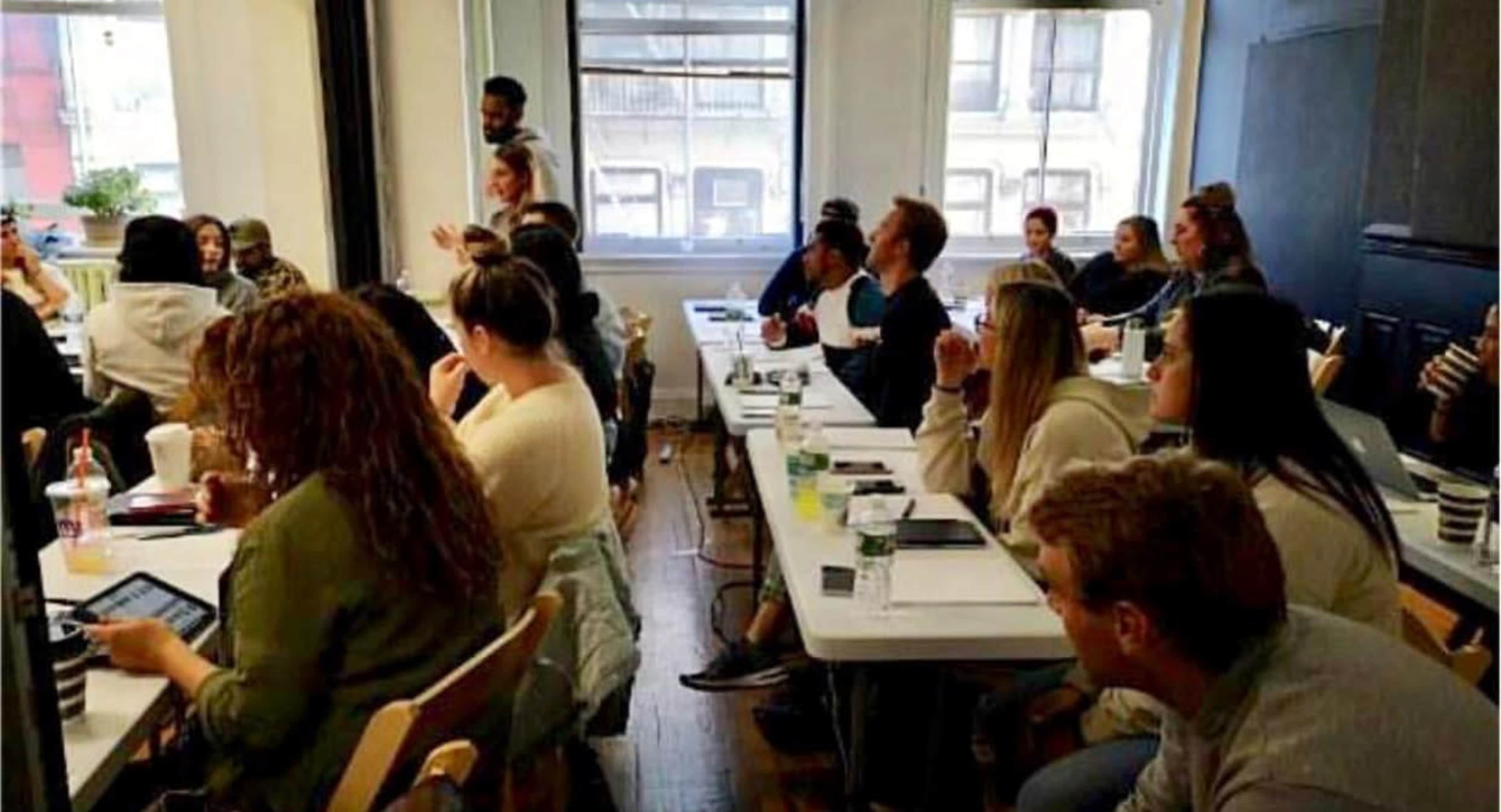 A group of individuals is seated at tables in a classroom setting, attentively listening to a speaker at the front of the room.
