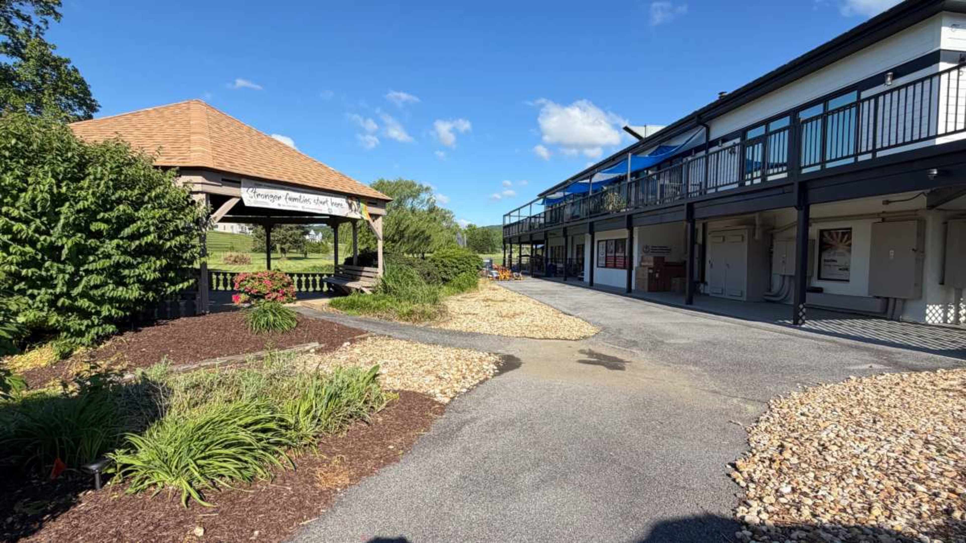 The image shows a pathway leading past a wooden gazebo and a building with multiple balconies against a clear blue sky.