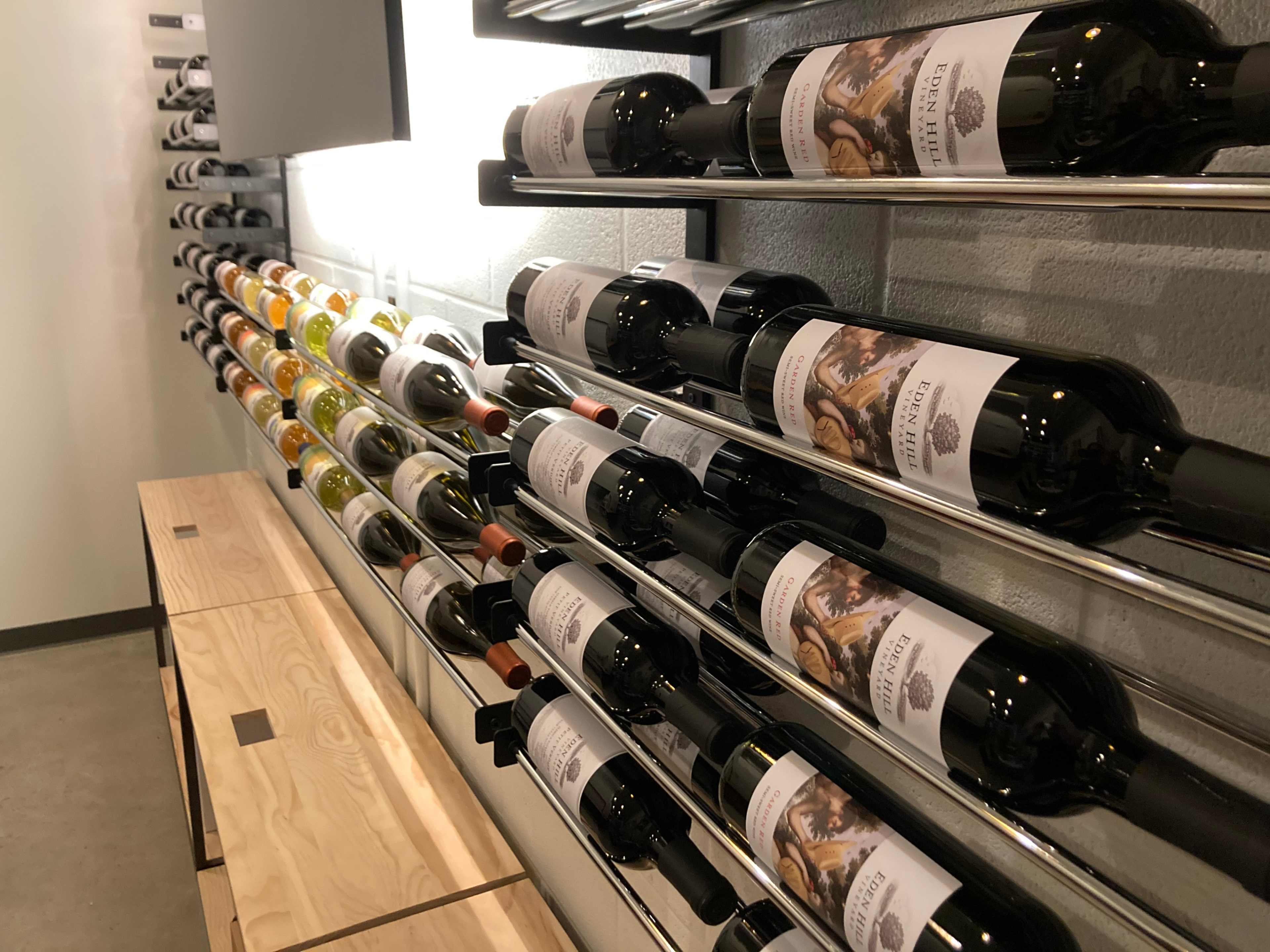 The image shows a display of wine bottles arranged on metal racks along a wall, with a wooden shelf below.