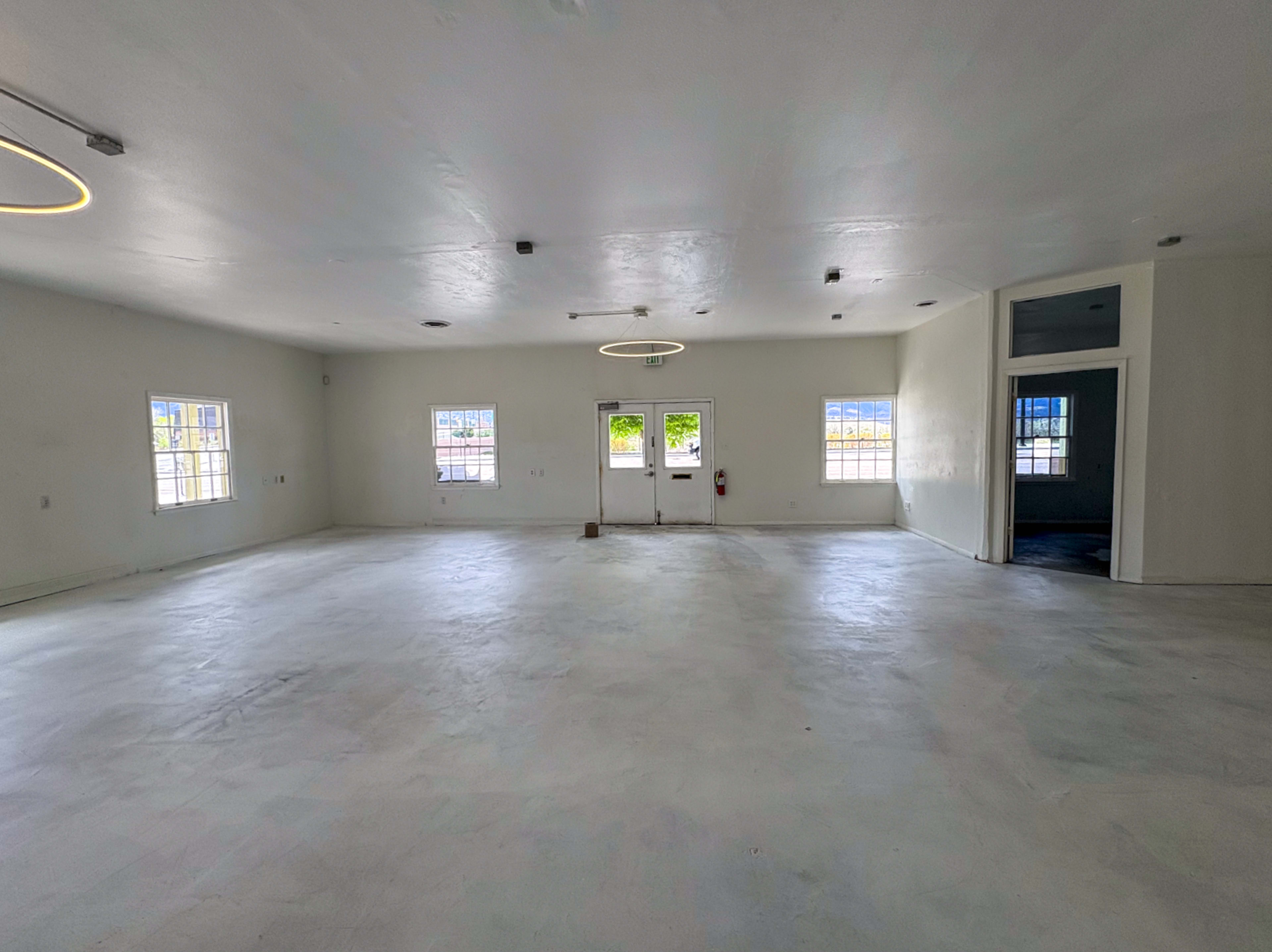 The image shows a spacious, empty room with white walls, a concrete floor, and large windows allowing natural light.