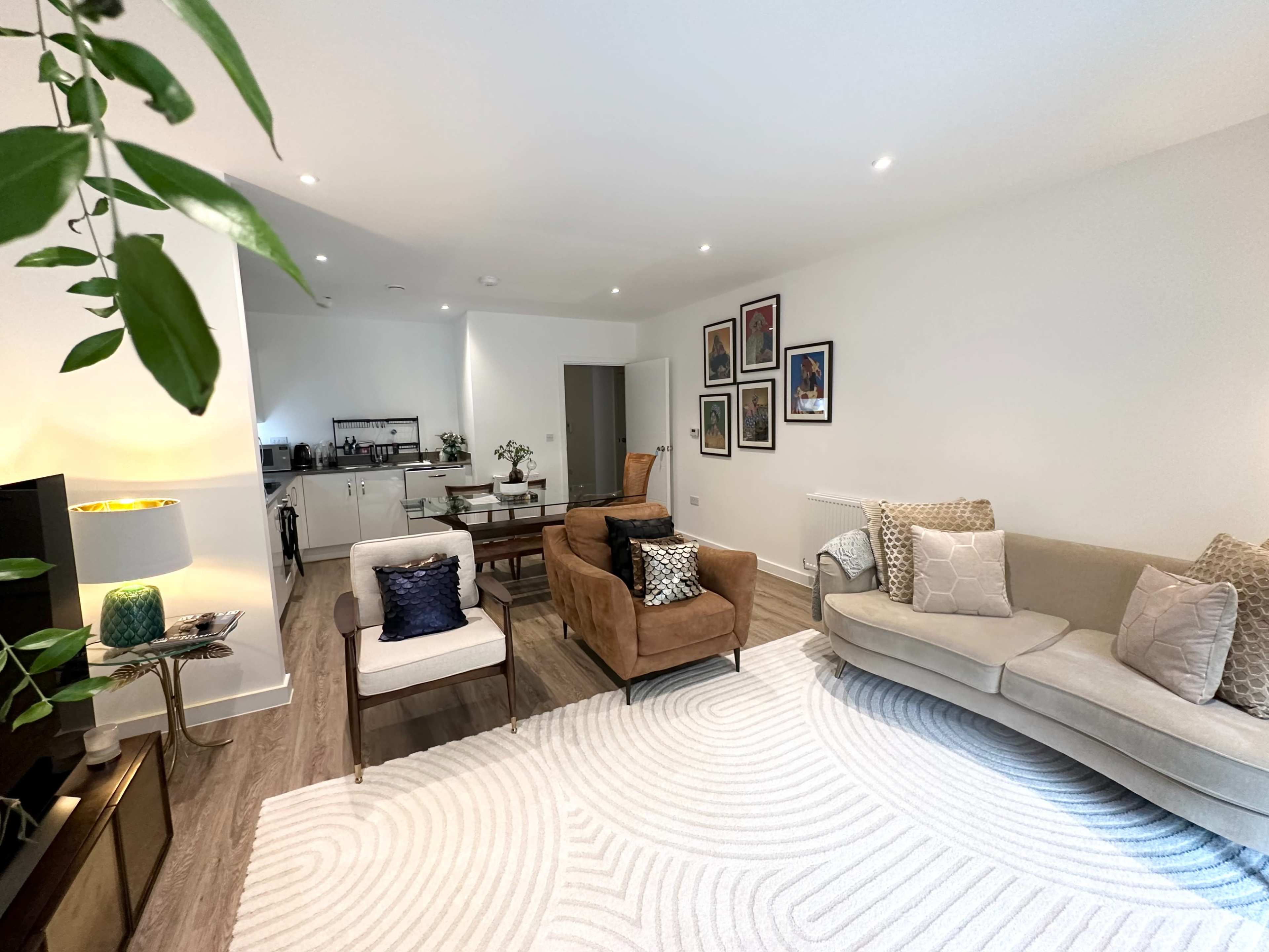 The living room features a light-colored sofa, two armchairs, a round rug, and framed artworks on the wall, with a small dining area visible in the background.