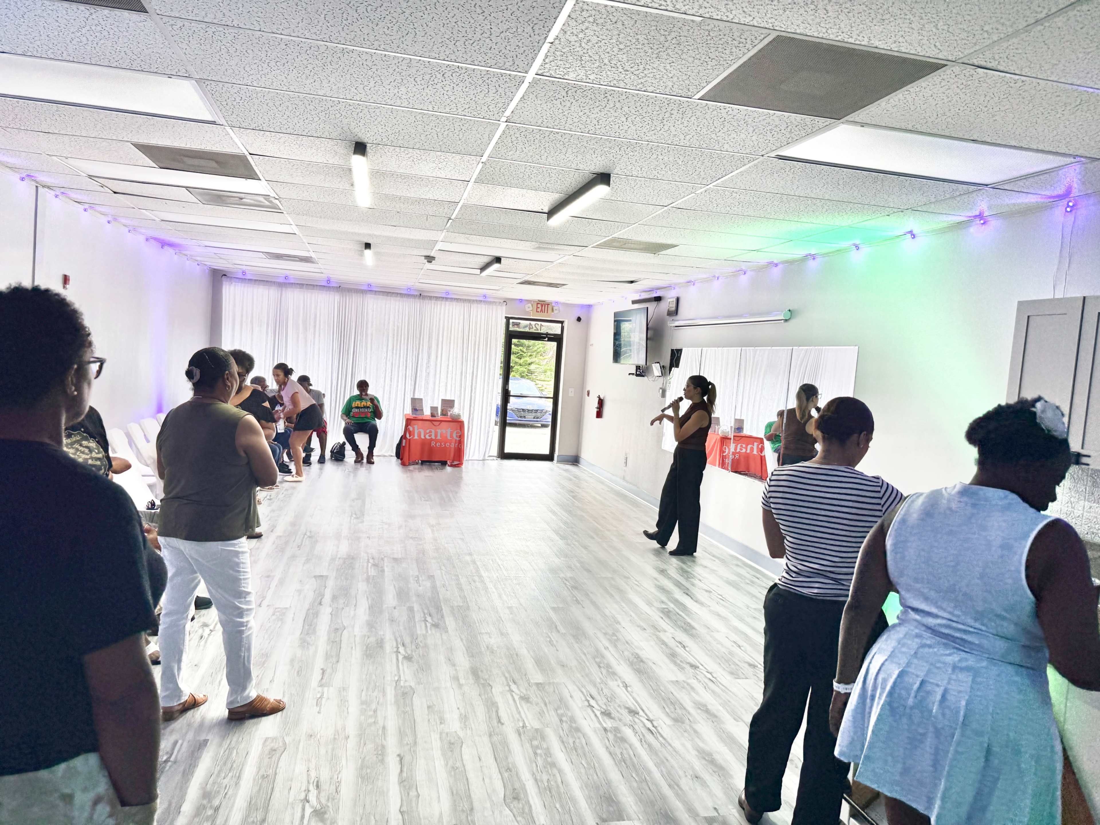 A group of people lines up inside a brightly lit room with a mirrored wall, while a person stands at the front, leading an activity or presentation.