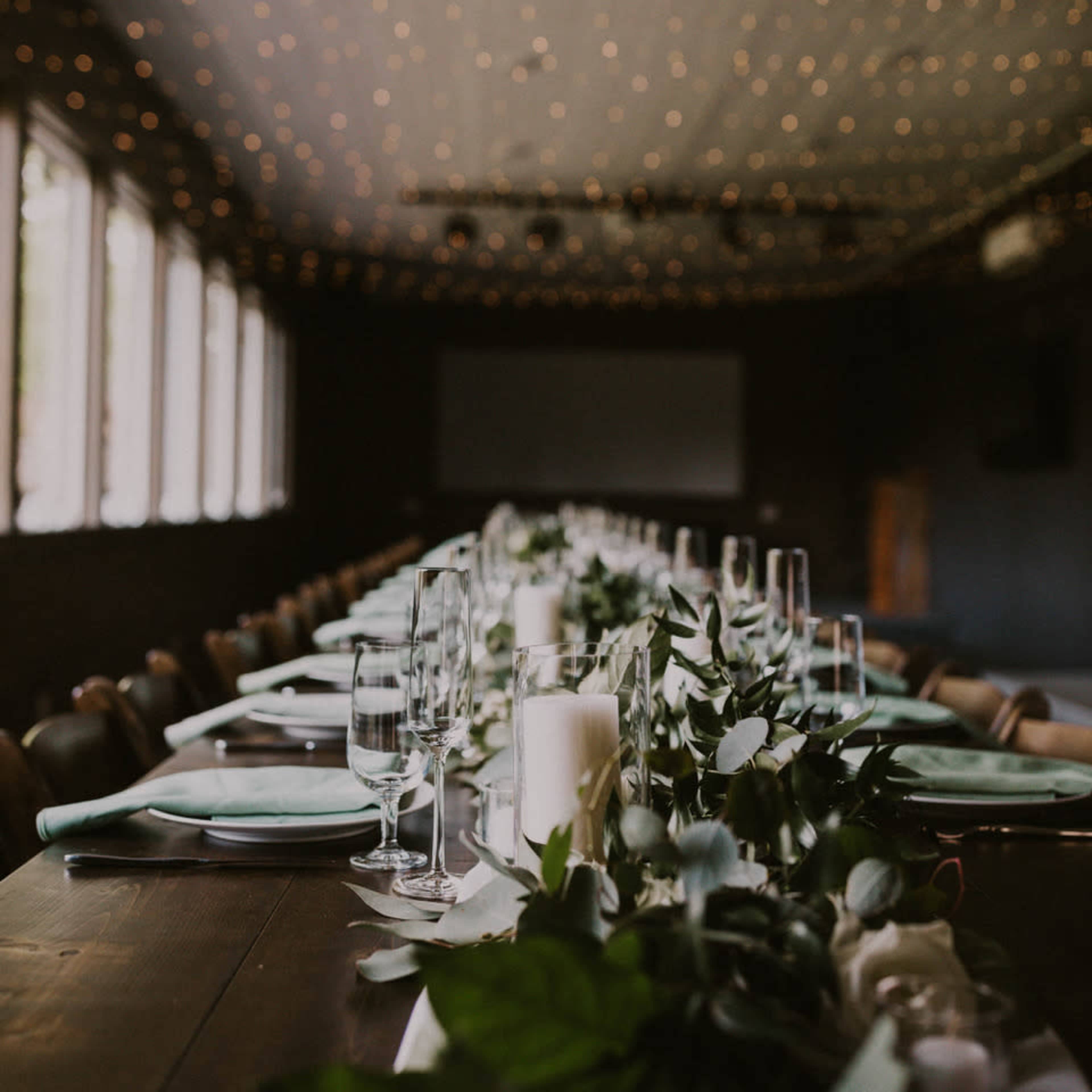 A long dining table is set with green napkins, glassware, and decorative foliage under softly lit string lights.