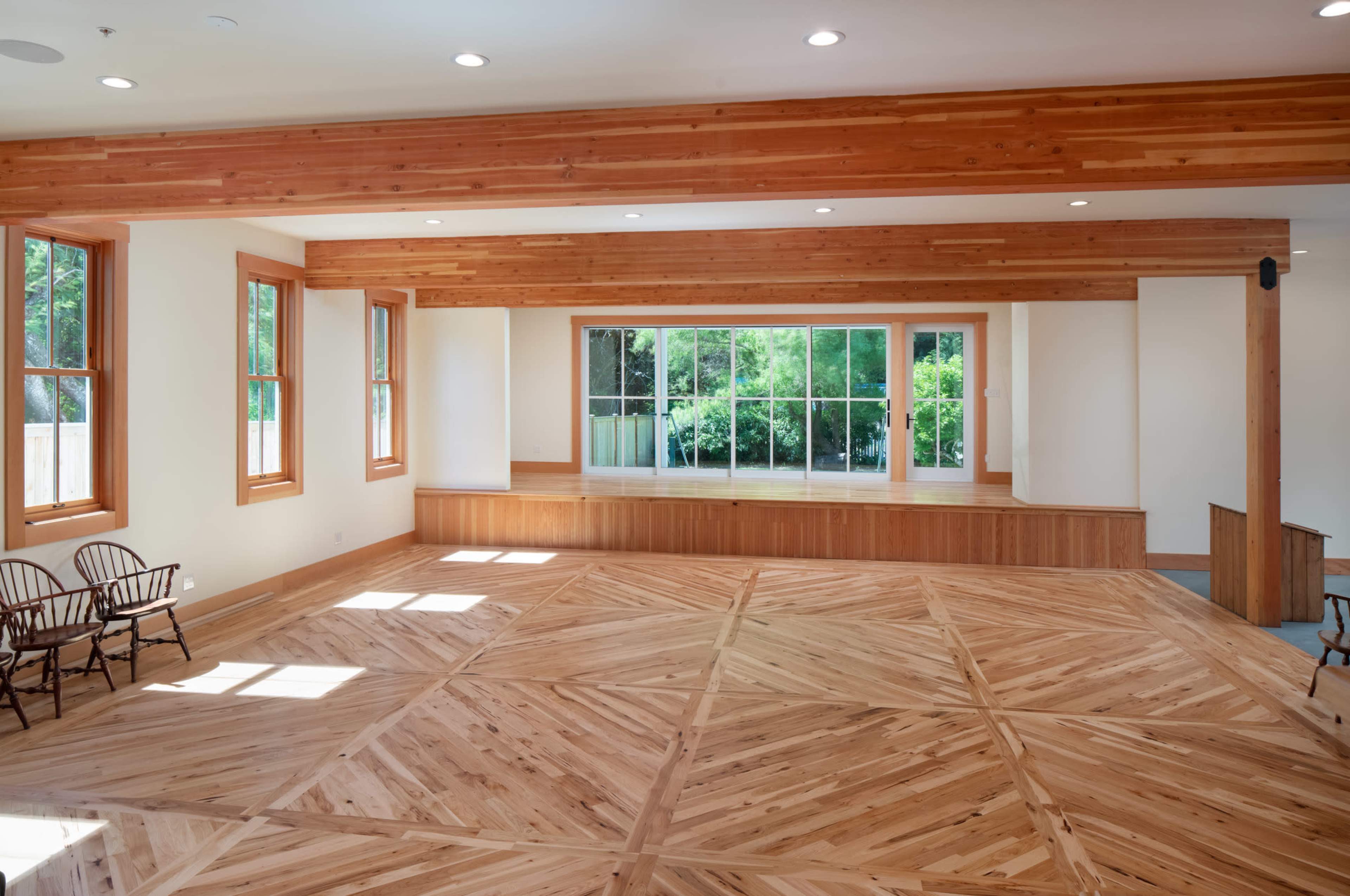 A spacious room with wooden flooring and large windows overlooking a green outdoor area.