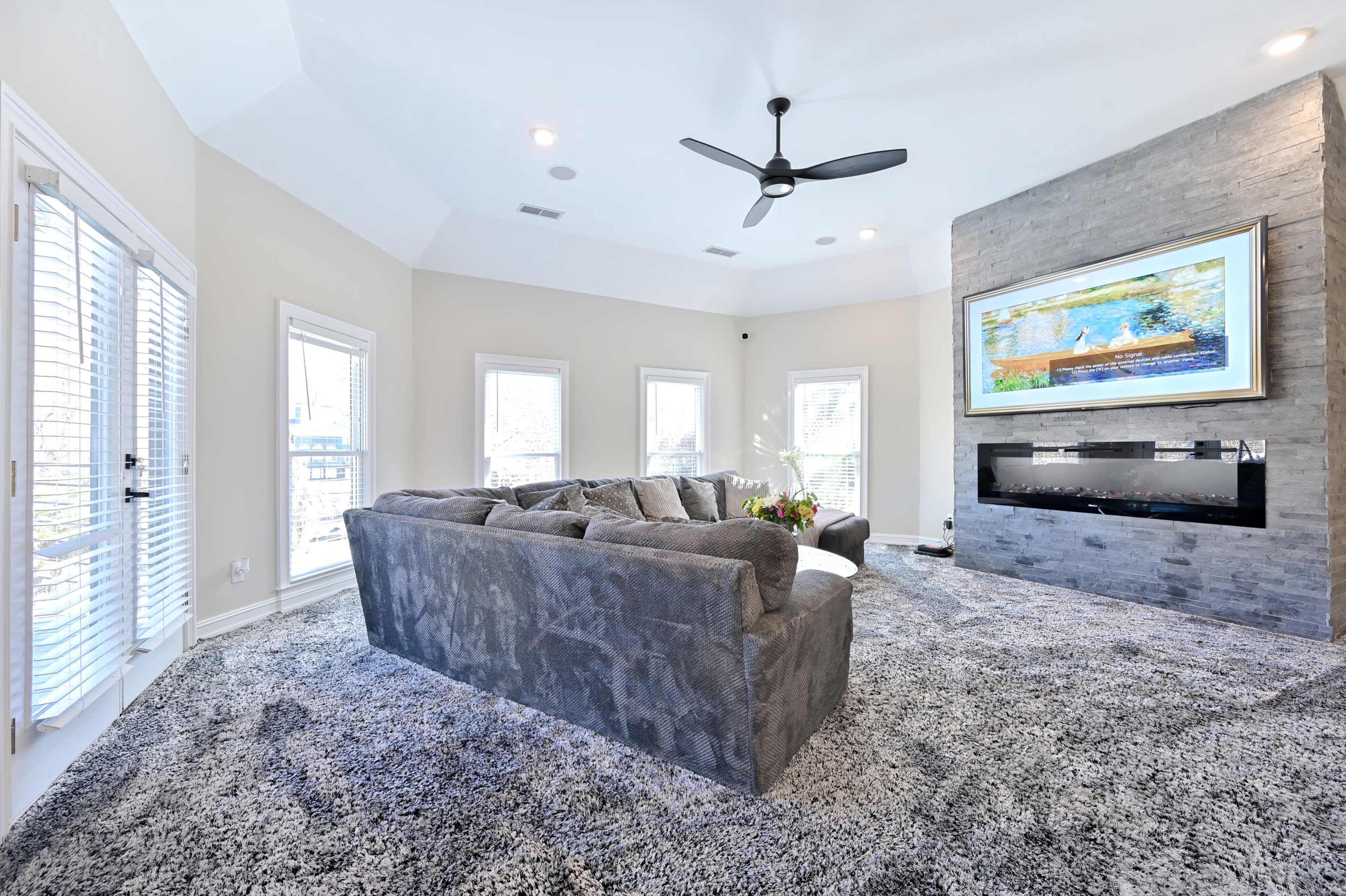 A cozy living room features a gray sectional sofa, a stone fireplace, and a large mounted television, with abundant natural light from surrounding windows.