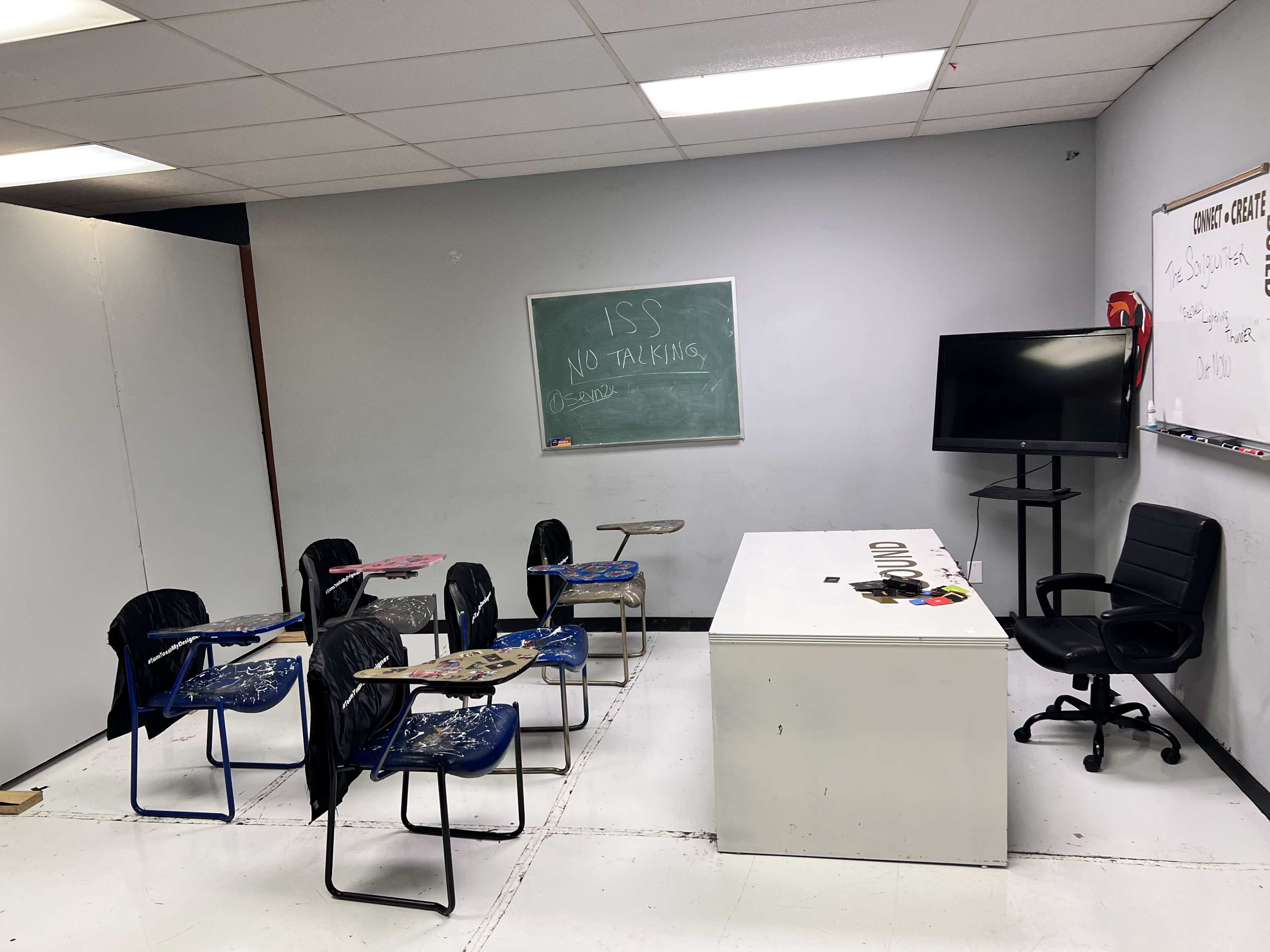 An empty classroom features several desks, a large white desk, a television, and a chalkboard with rules written on it.