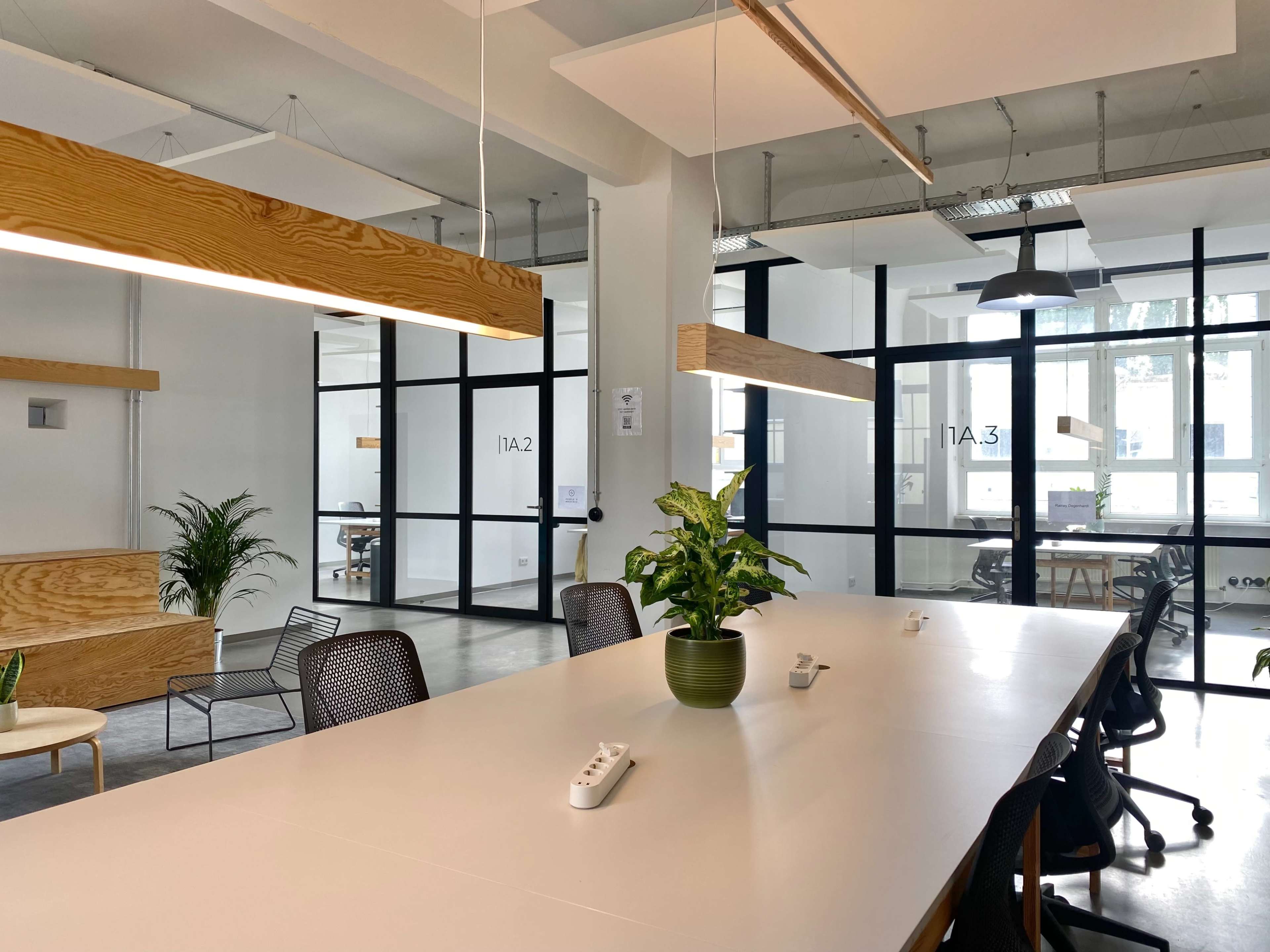 A modern office space features a long white table surrounded by black chairs, with wooden light fixtures and plants, adjacent to glass-walled meeting rooms.