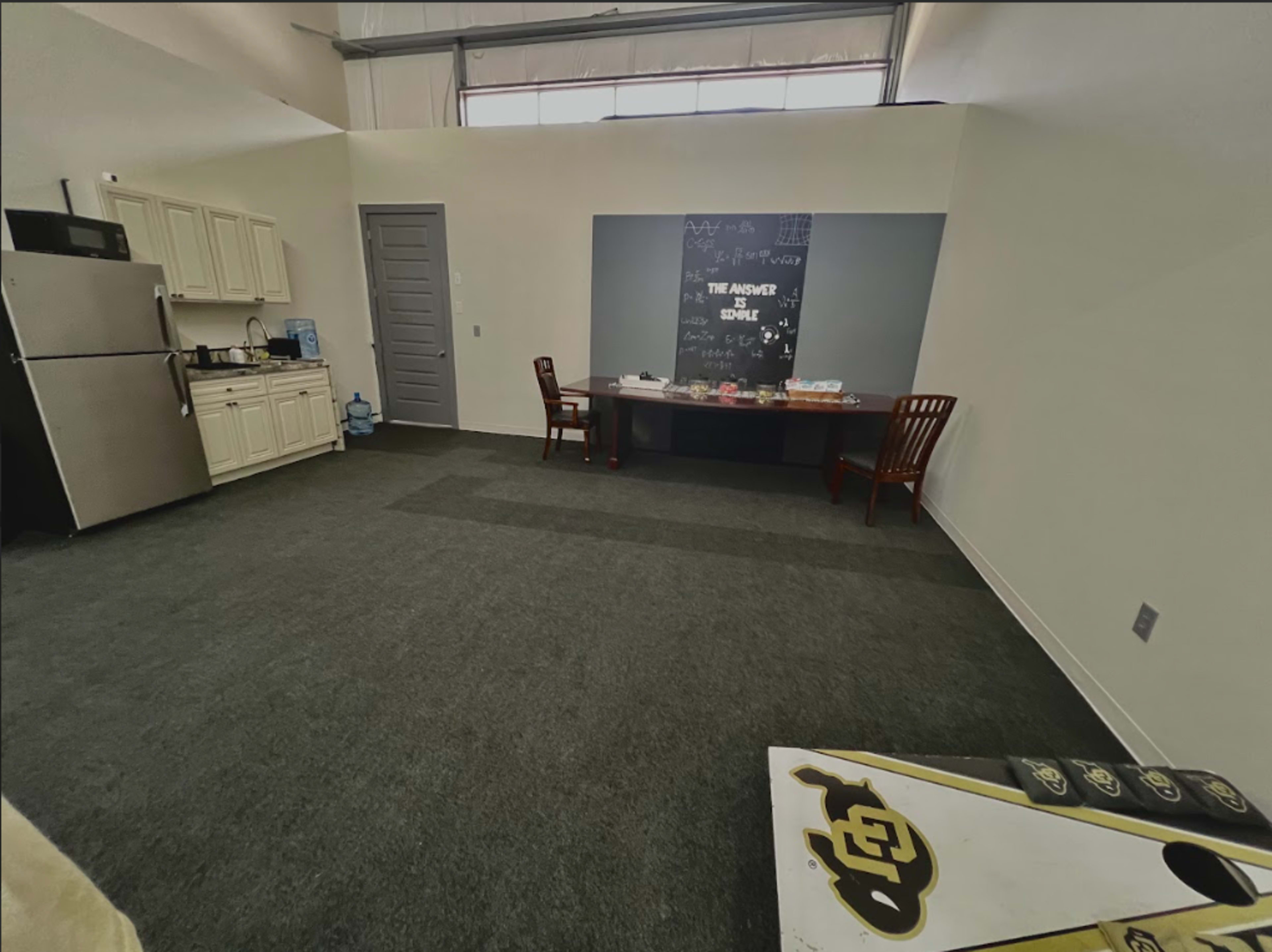 The image shows a sparsely furnished room with a kitchenette, two wooden chairs, and a table against a wall featuring a chalkboard.