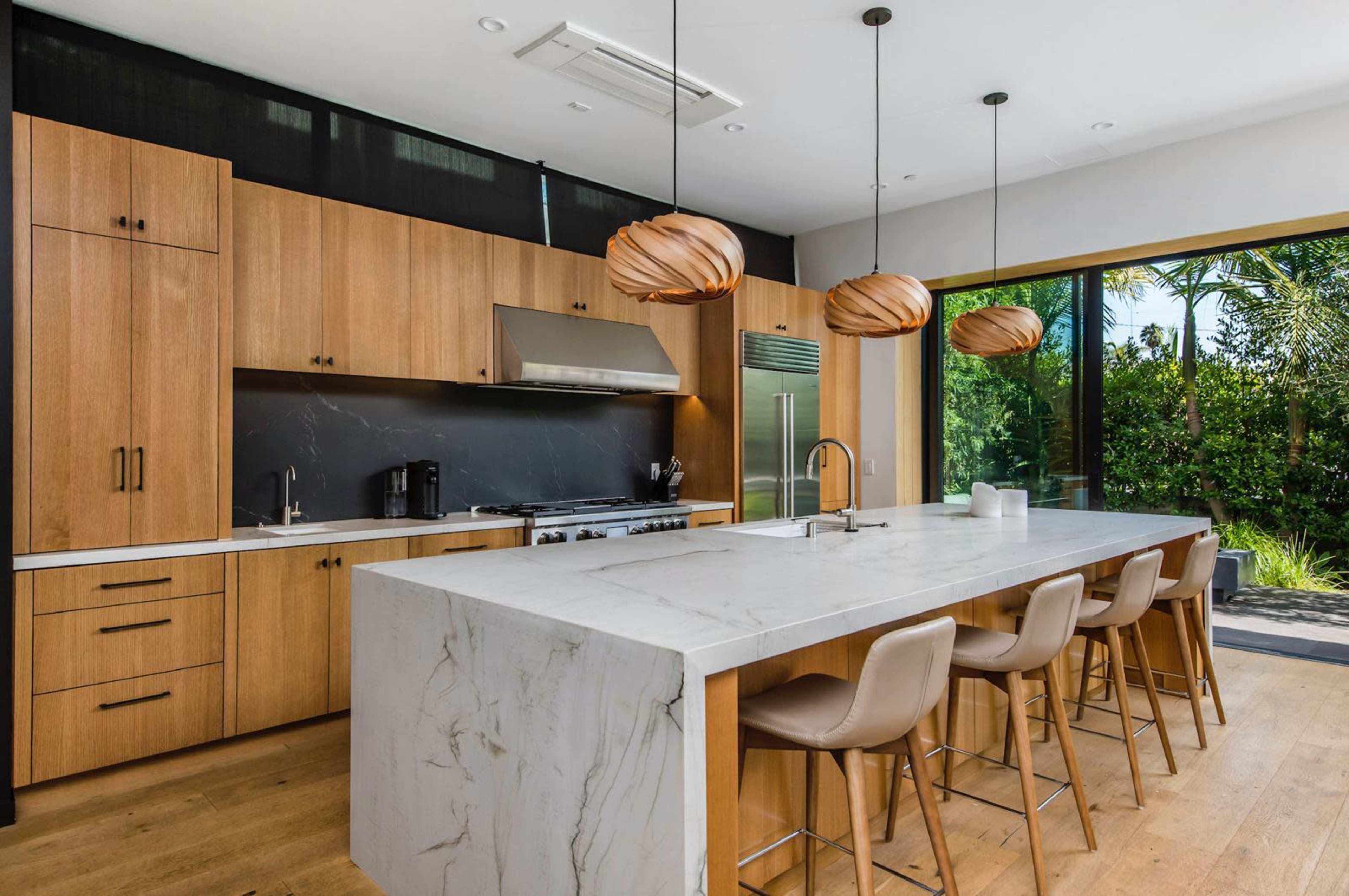 The image shows a modern kitchen with wooden cabinetry, a marble island with seating, and large windows overlooking a garden.