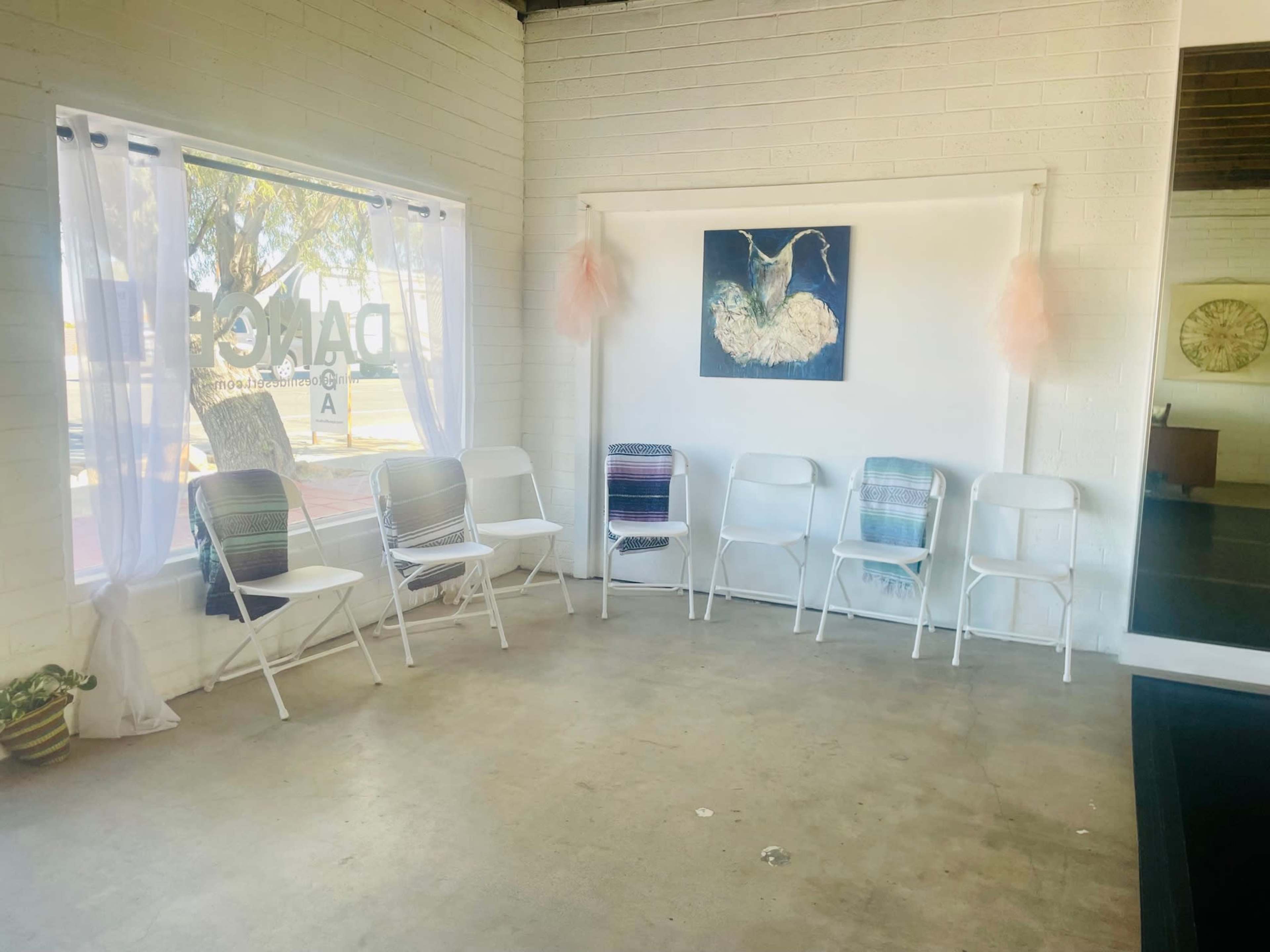 The image shows a simple interior space with white walls, a window, and a row of white chairs arranged in a circle under a blue artwork.