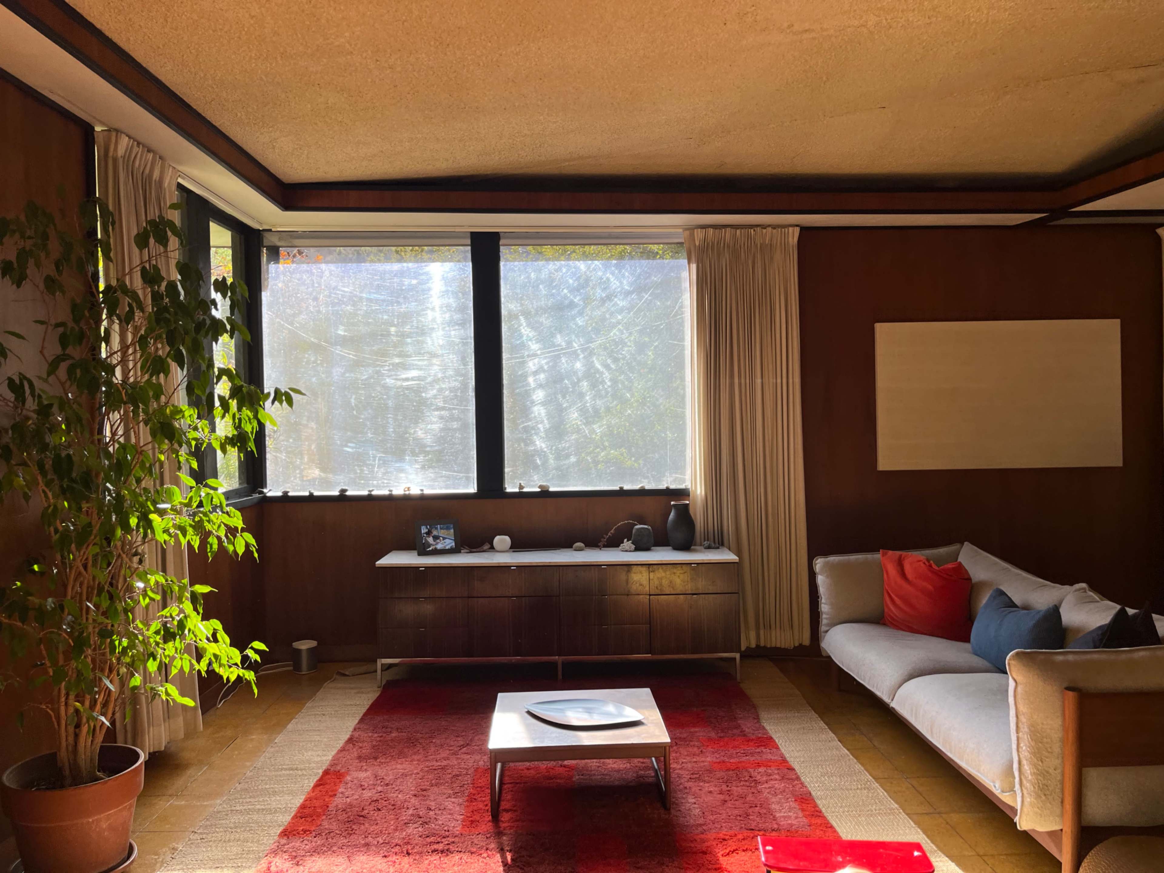 The image shows a mid-century modern living room with a sofa, a coffee table, a potted plant, and large windows allowing natural light to enter.