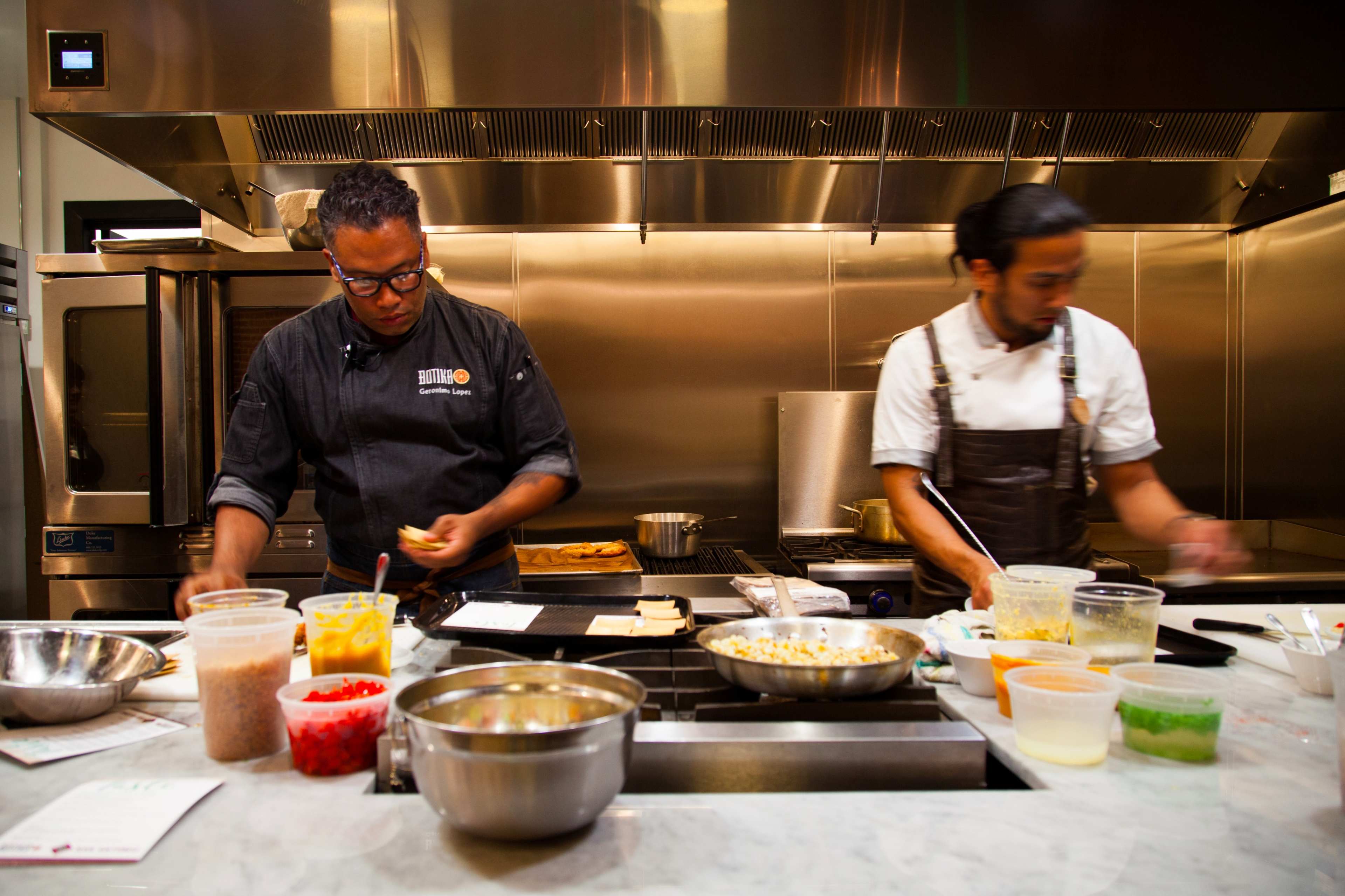Two chefs are preparing food in a modern kitchen, surrounded by various containers and utensils on a marble countertop.
