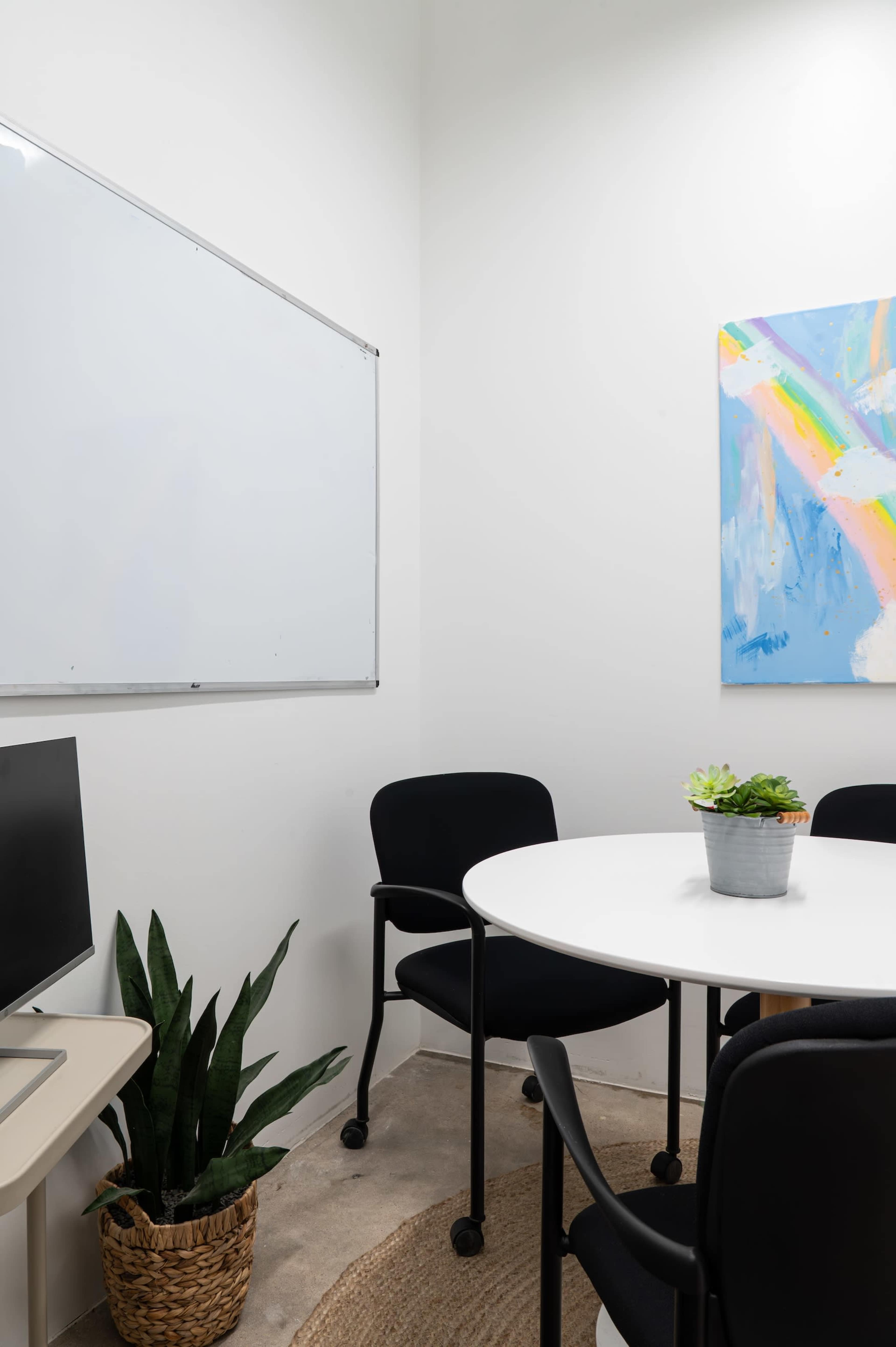 A small meeting room features a whiteboard, a round table with chairs, a computer desk, and a potted plant.