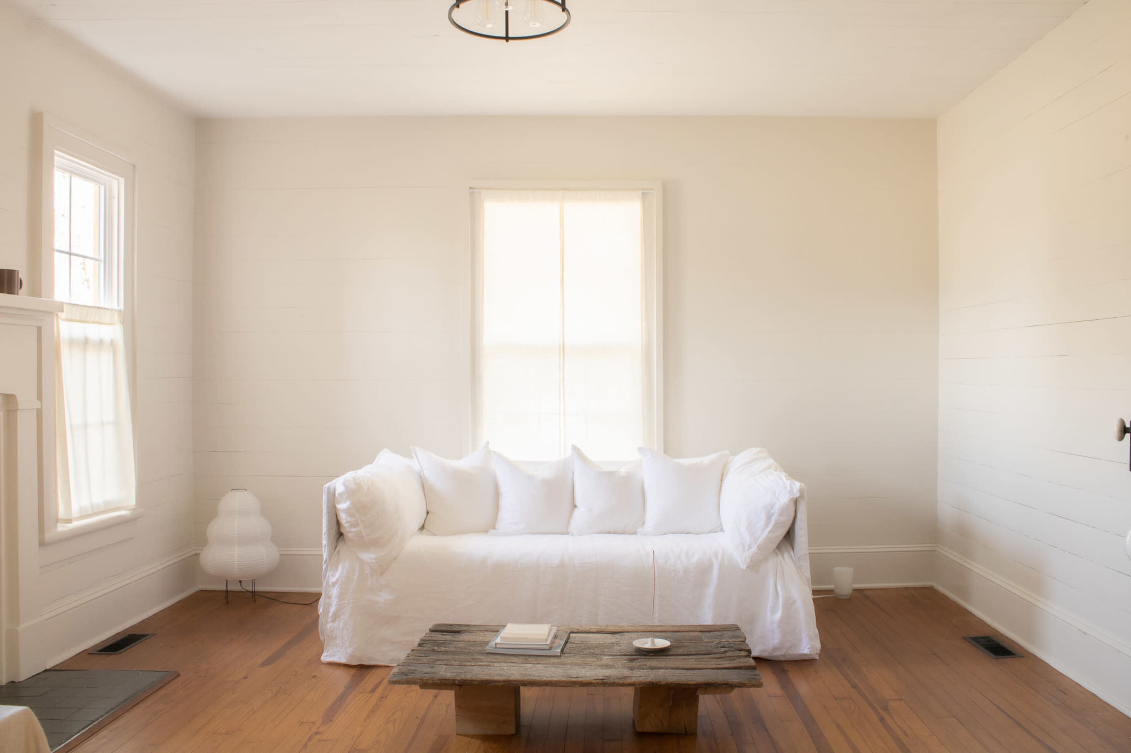 A bright, minimalistic living room features a white sofa with multiple cushions, a wooden coffee table, and wooden flooring.