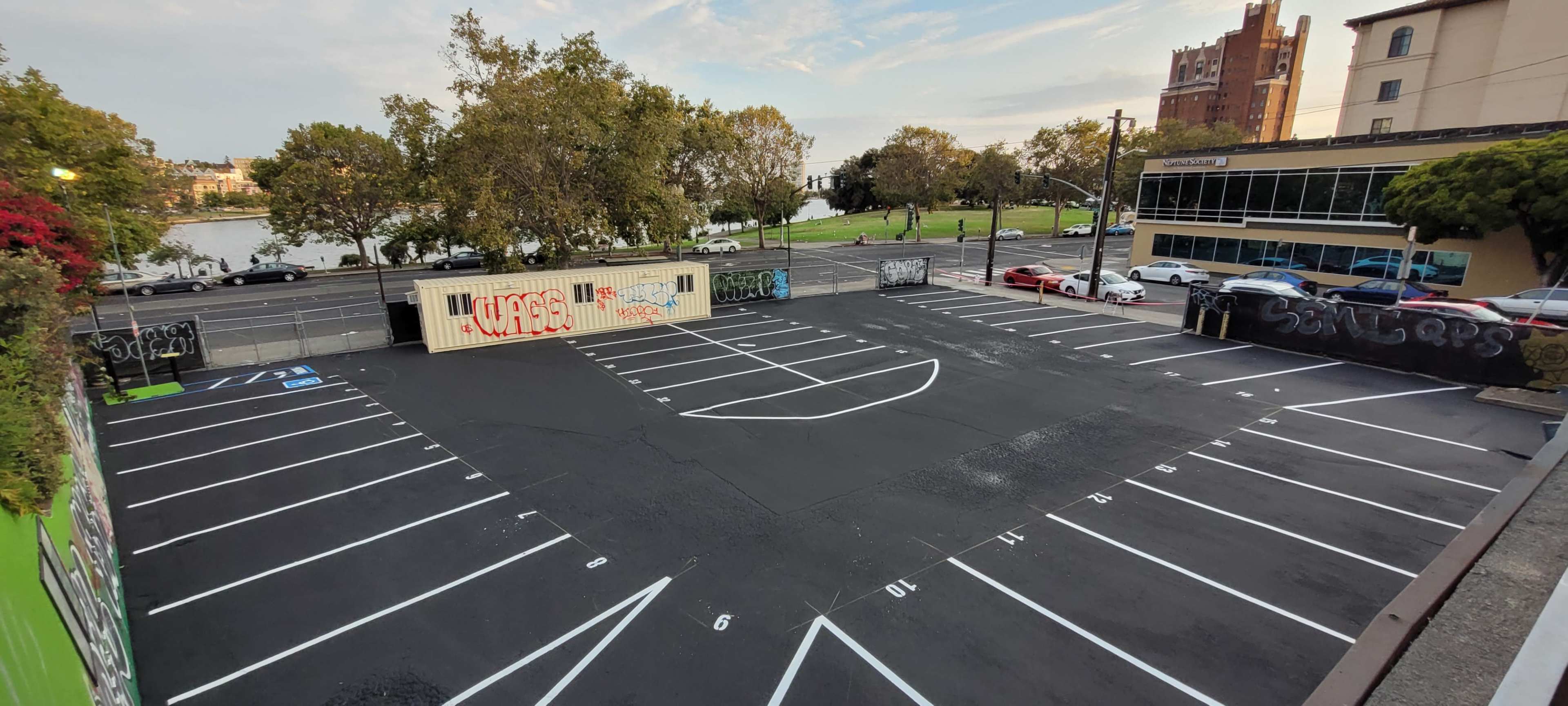 An empty parking lot with freshly painted white lines and a shipping container nearby, set against a backdrop of trees and a building.