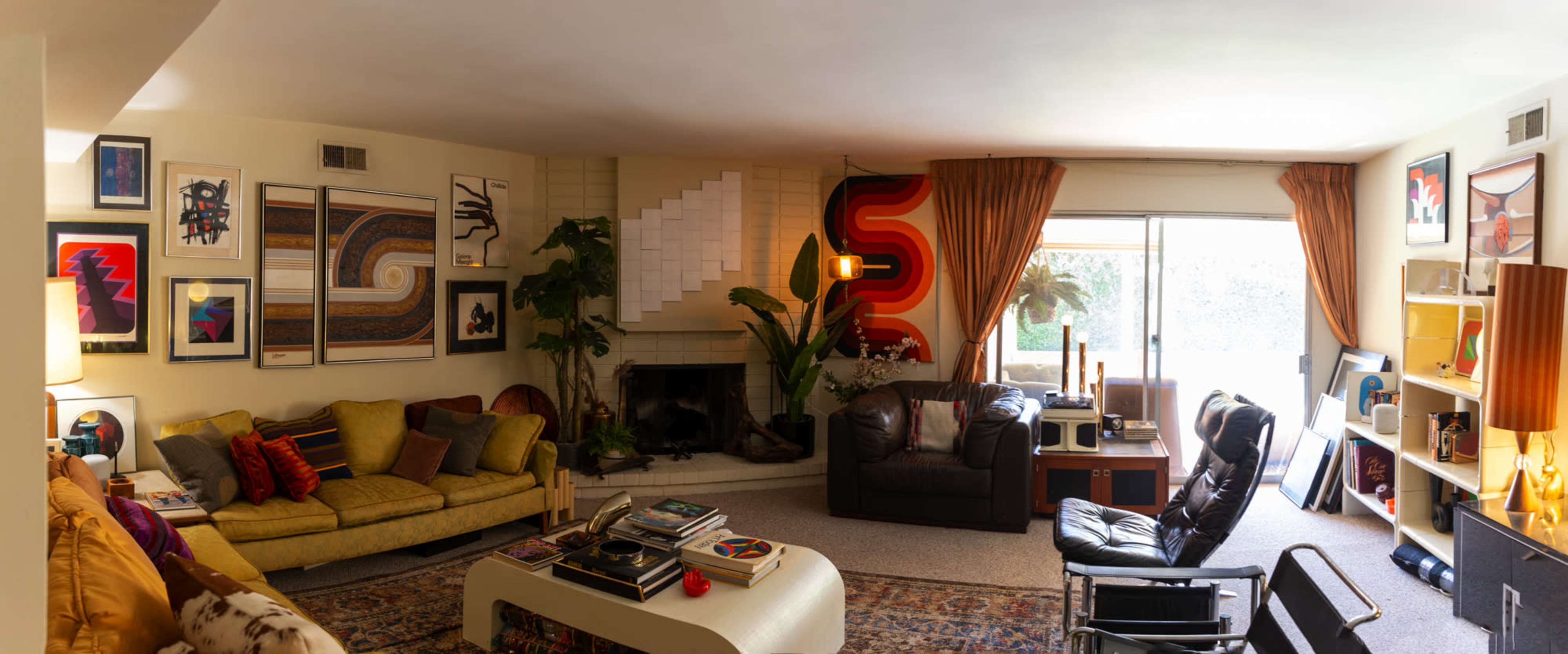 The image shows a spacious living room featuring colorful artwork, a green sofa, a black leather chair, and large windows letting in natural light.
