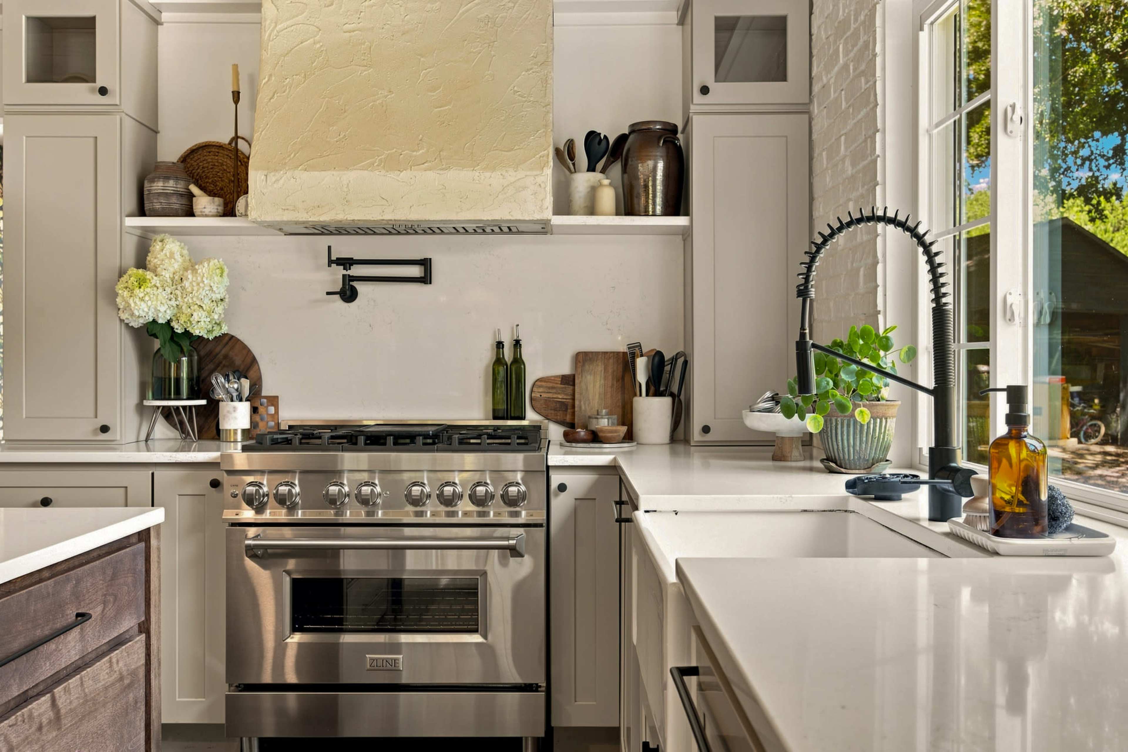 The image shows a modern kitchen with a stainless steel oven, white cabinetry, and a large window that provides natural light.