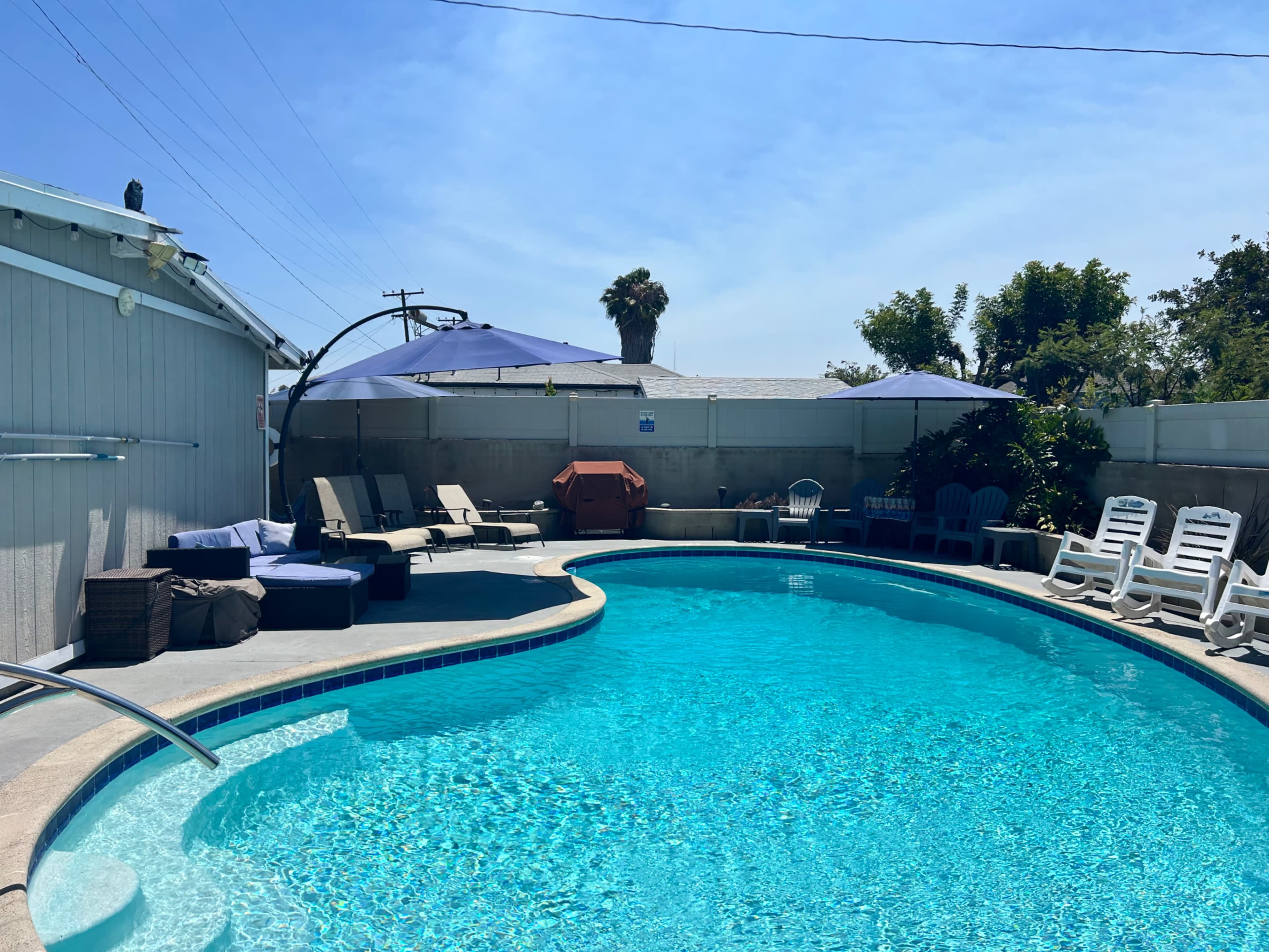 The image shows a turquoise swimming pool surrounded by lounge chairs and umbrellas in a sunny outdoor setting.
