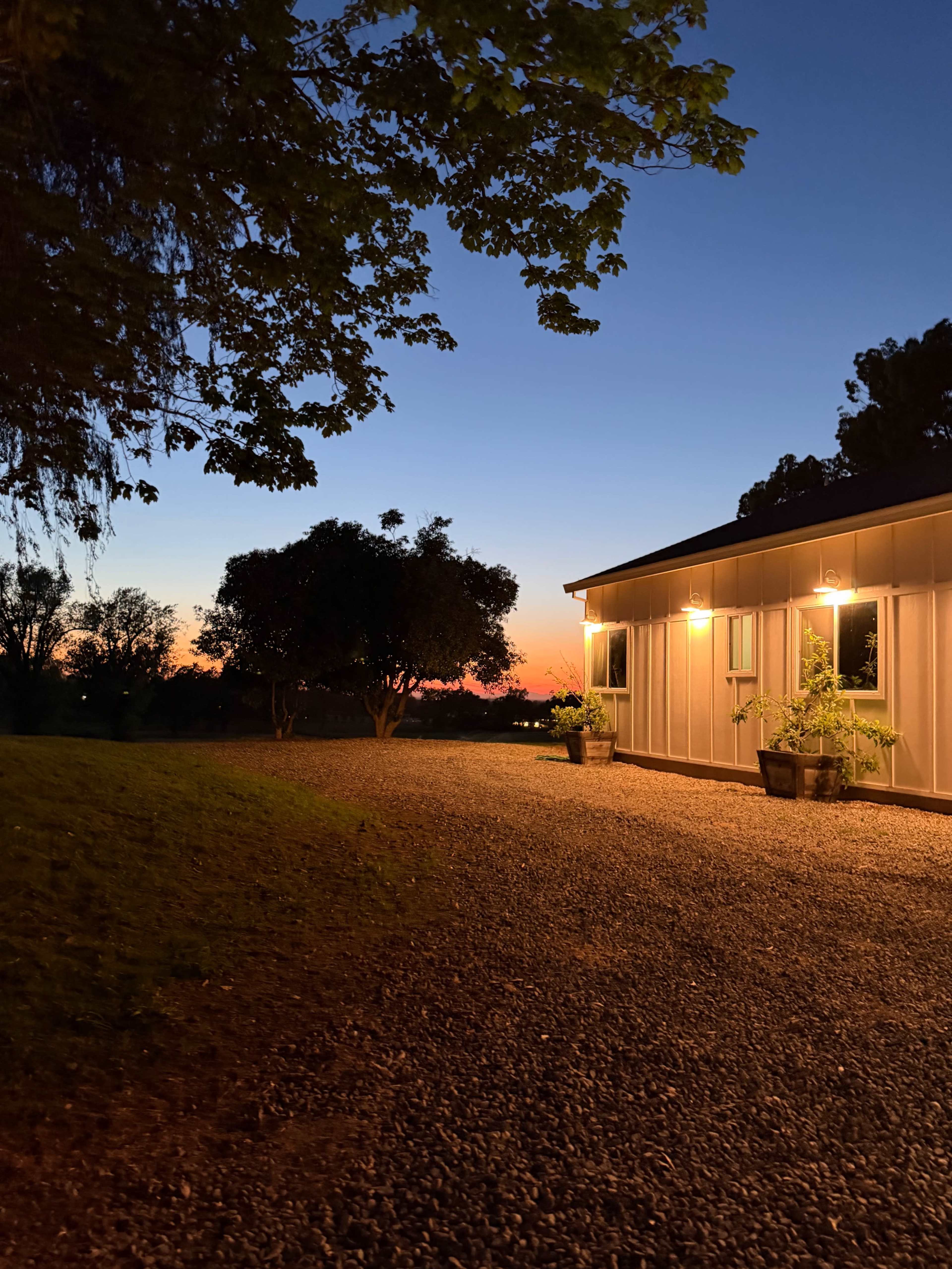A gravel path leads to a house with illuminated windows, framed by trees and a vivid sunset sky.