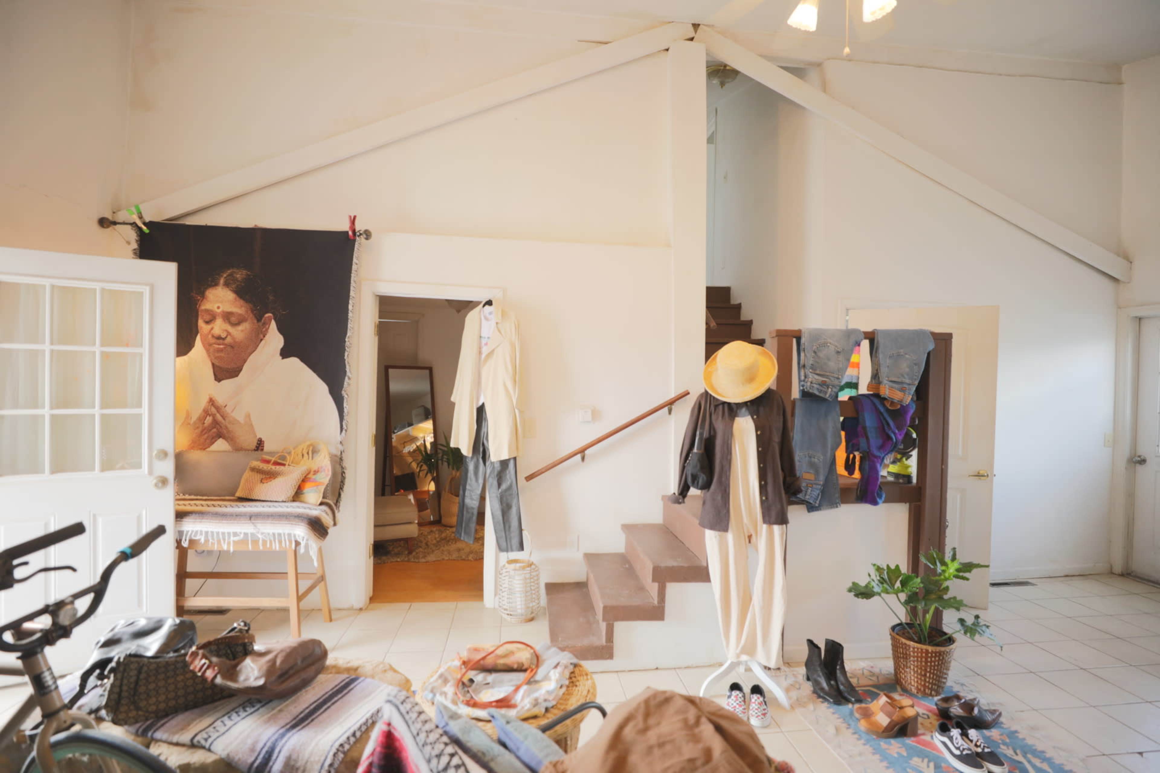 The image shows a spacious room featuring a staircase, clothing hanging on a rack and walls, and a large fabric print of a woman using a laptop.