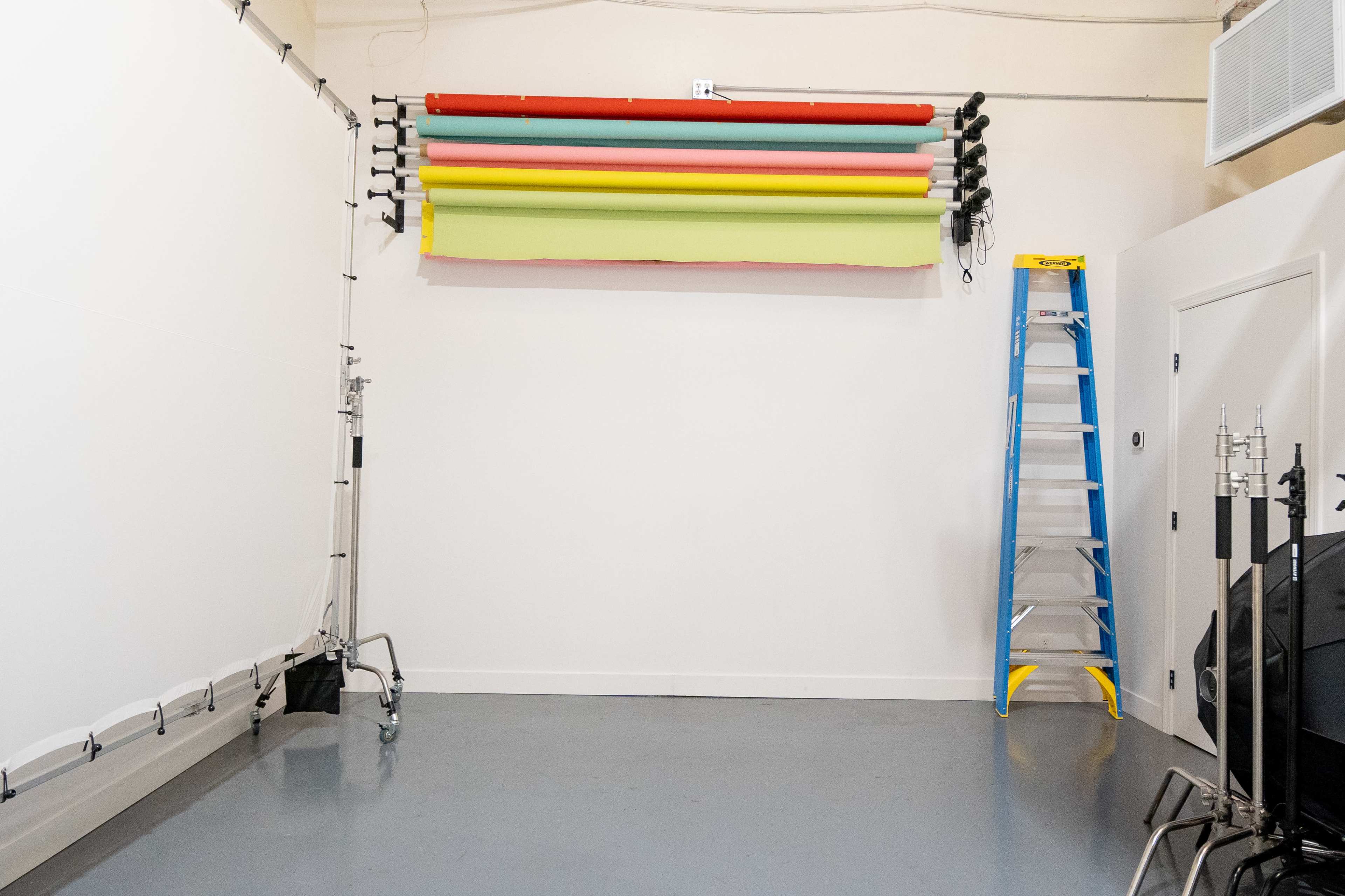 A ladder stands next to a wall-mounted backdrop system with various colored rolls in an empty studio space.