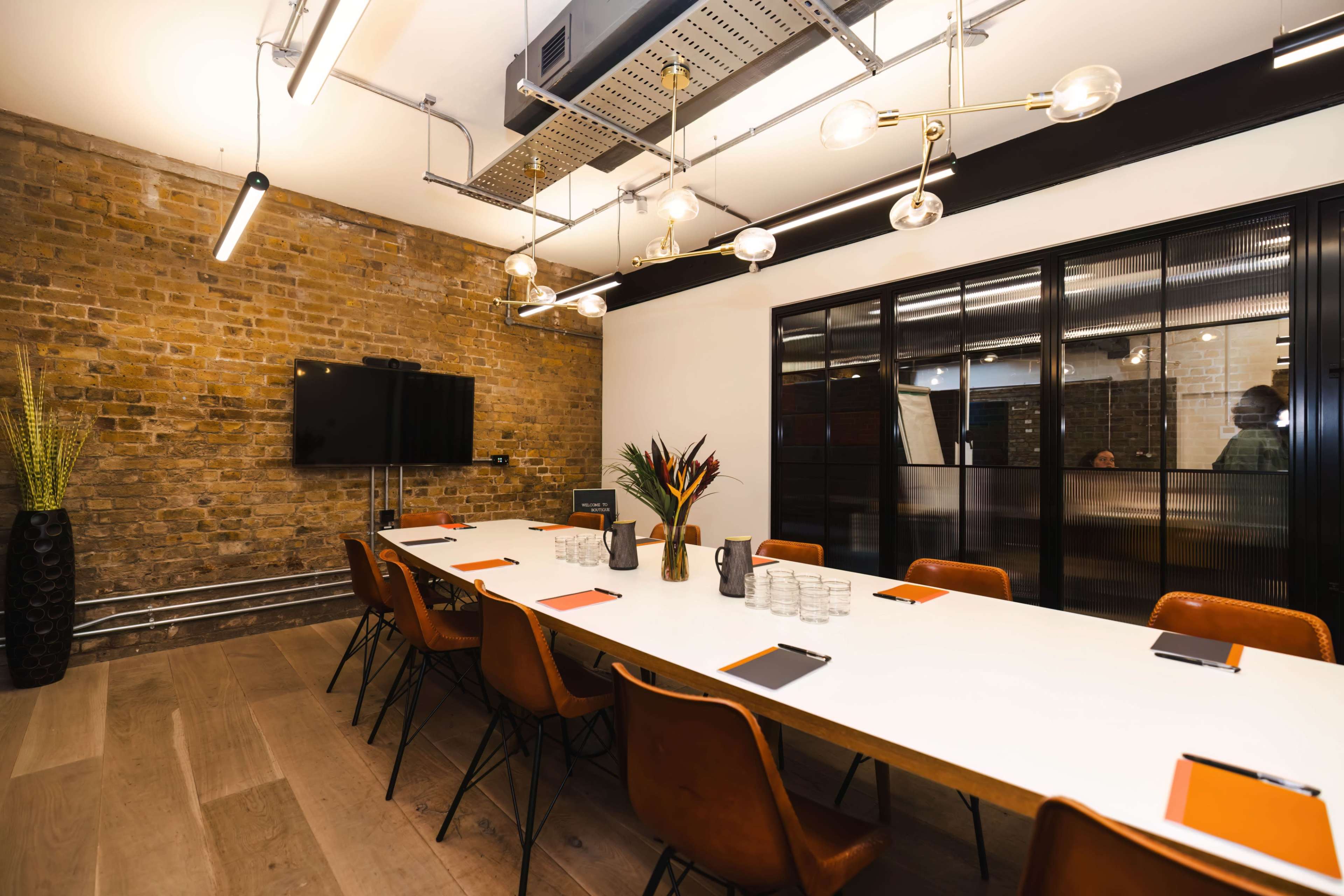 A long, white table with brown chairs is set for a meeting in a room featuring exposed brick walls and modern lighting.