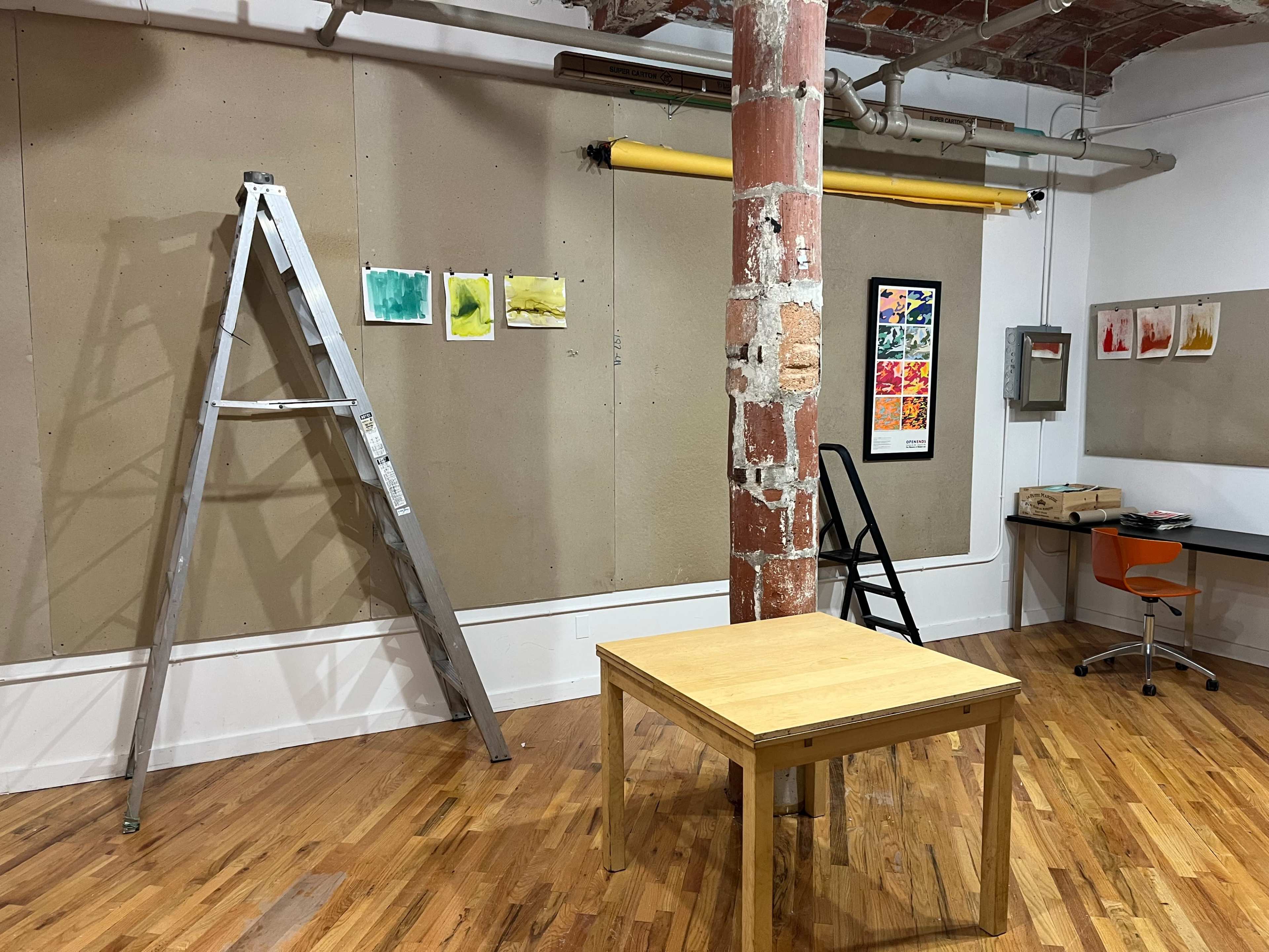 An empty art studio with a wooden table, a ladder, and several colorful paintings displayed on the walls.