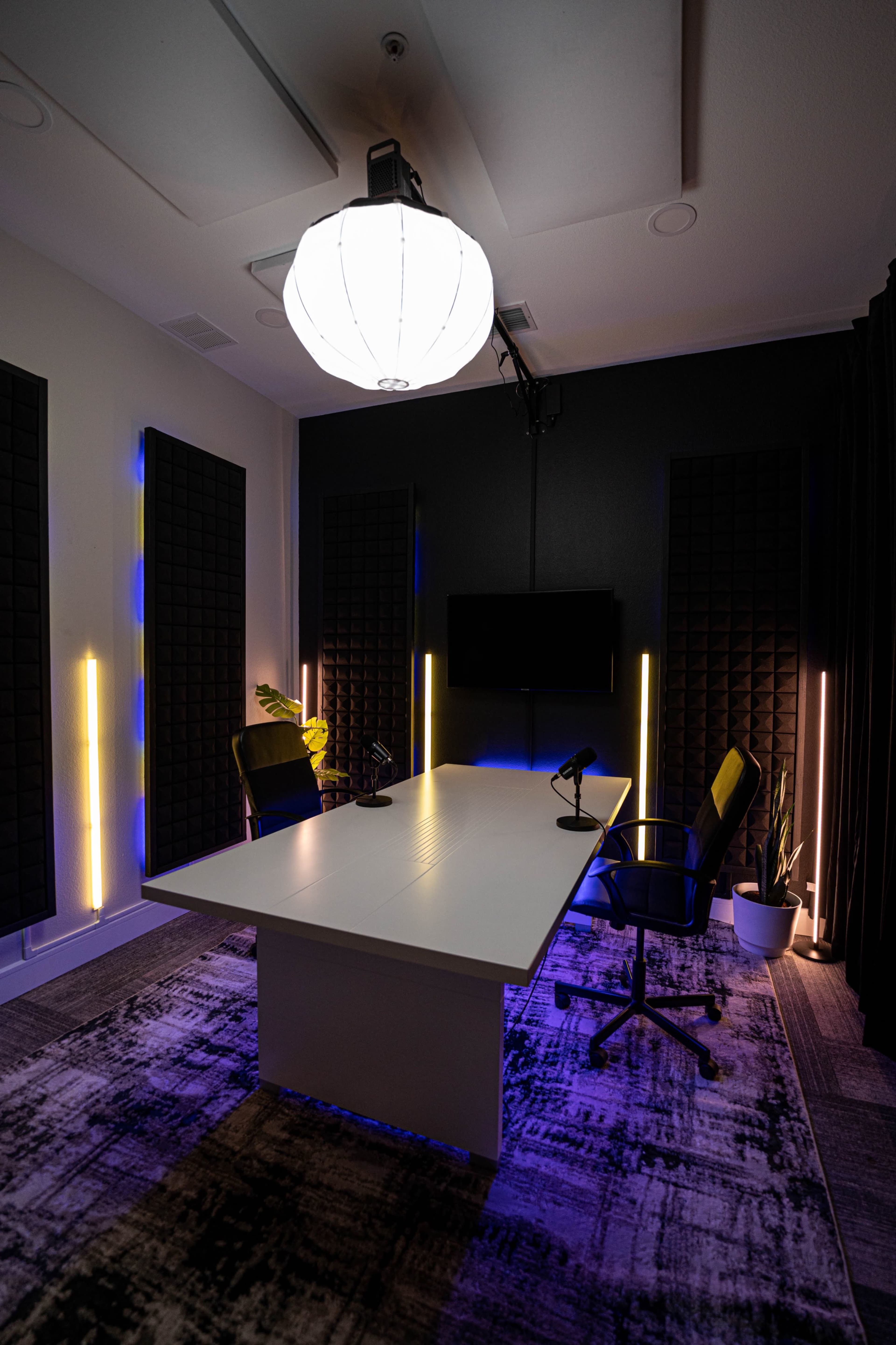 A modern conference room features a central table with microphones, surrounded by two chairs, and illuminated by soft lighting with acoustic panels on the walls.