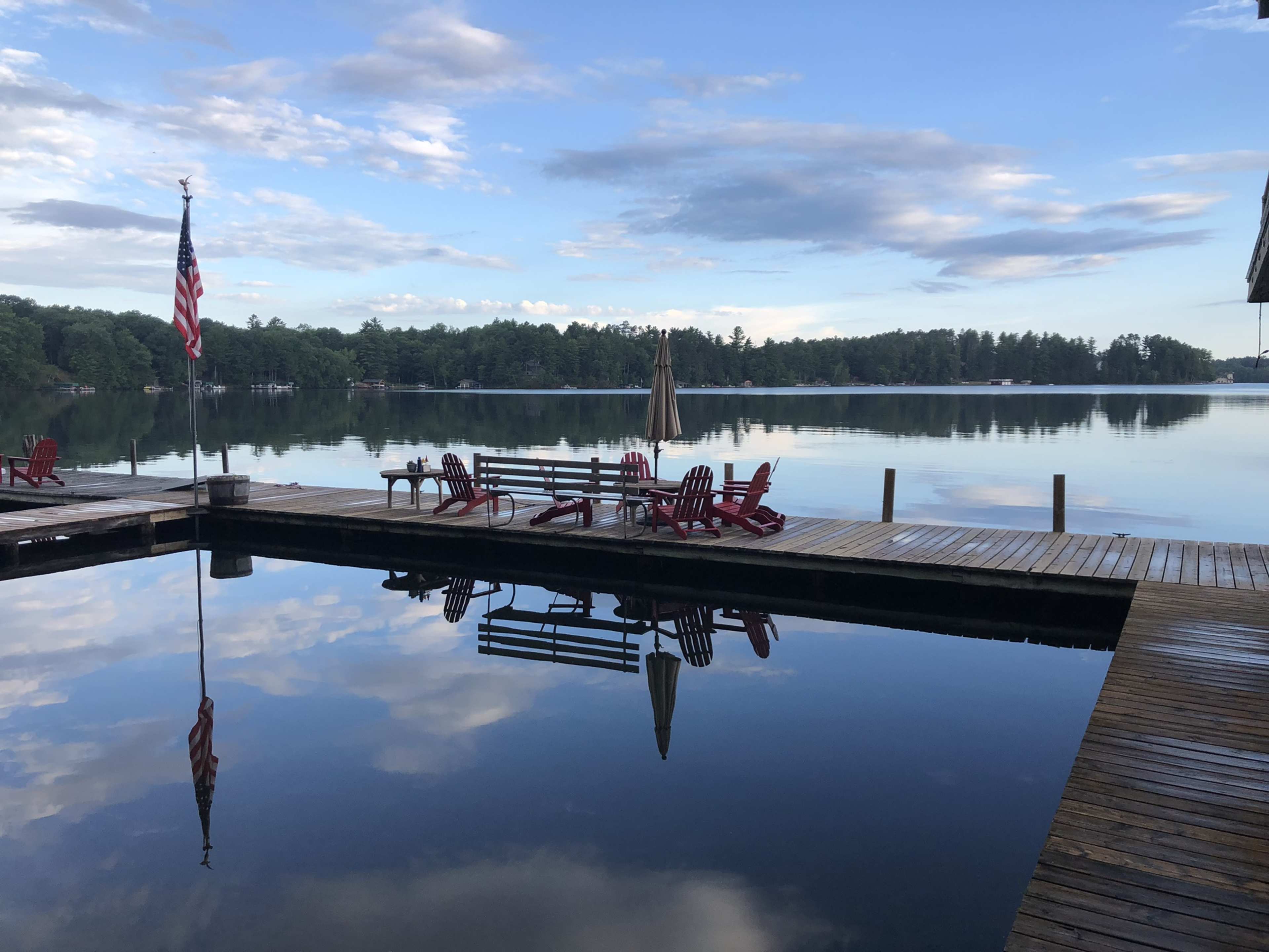 The image shows a calm lake with a wooden dock, featuring red Adirondack chairs and an American flag reflecting in the water.
