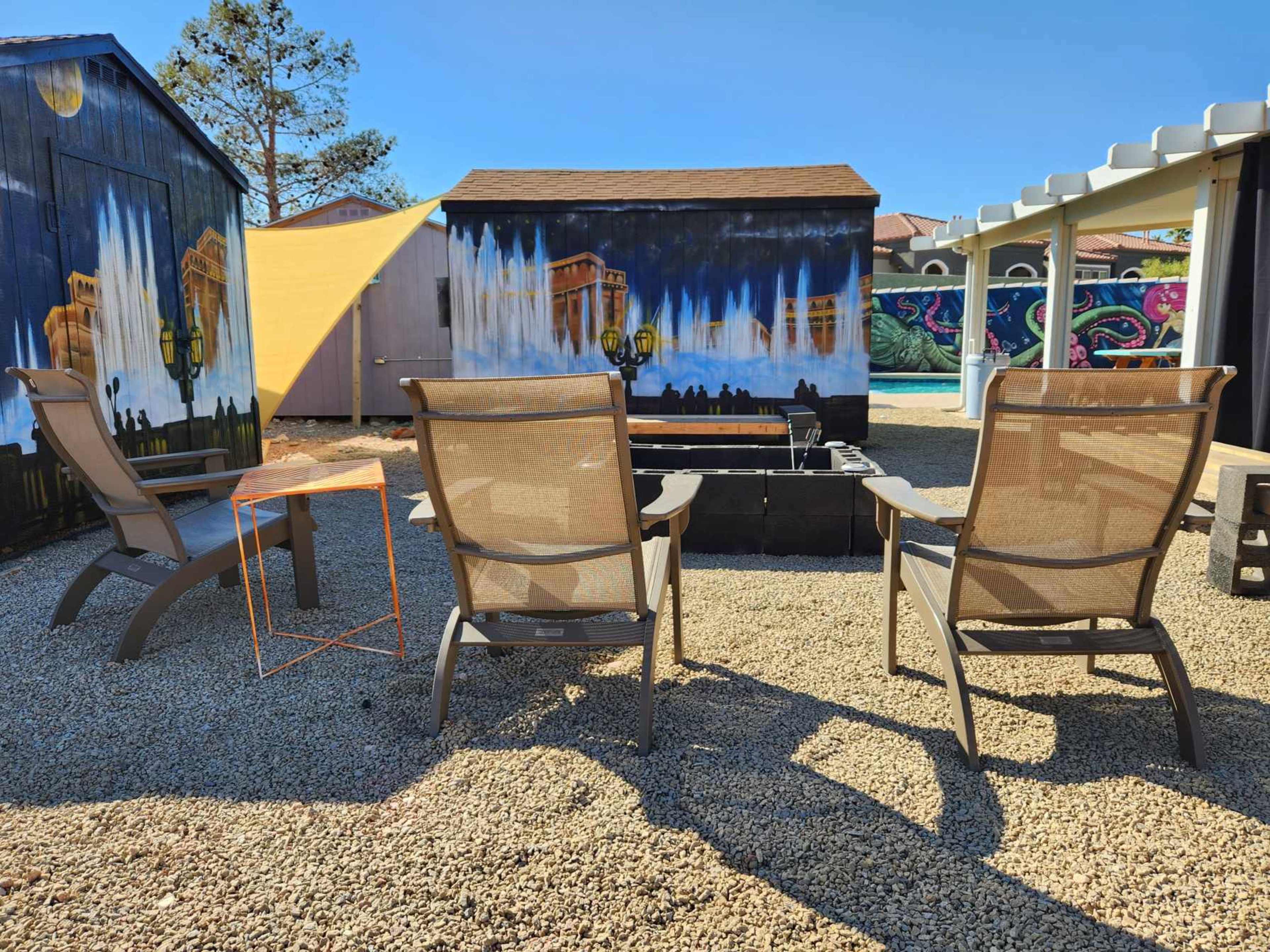 Three lounge chairs are arranged around a small table in a gravel courtyard beside colorful murals on nearby walls.