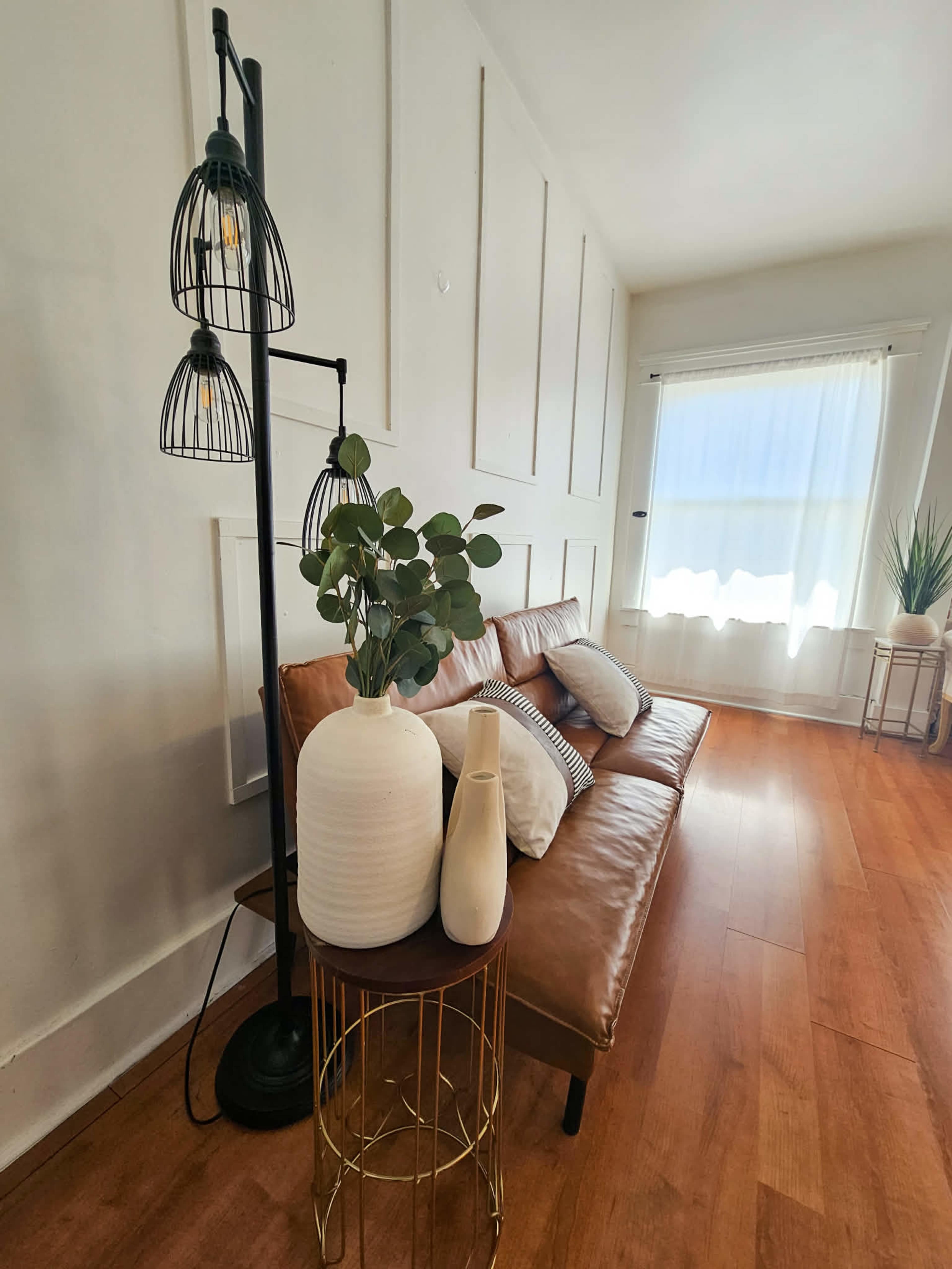 The image shows a well-lit living room featuring a brown leather sofa, decorative pillows, a white vase with greenery, and a round side table beside a large window.