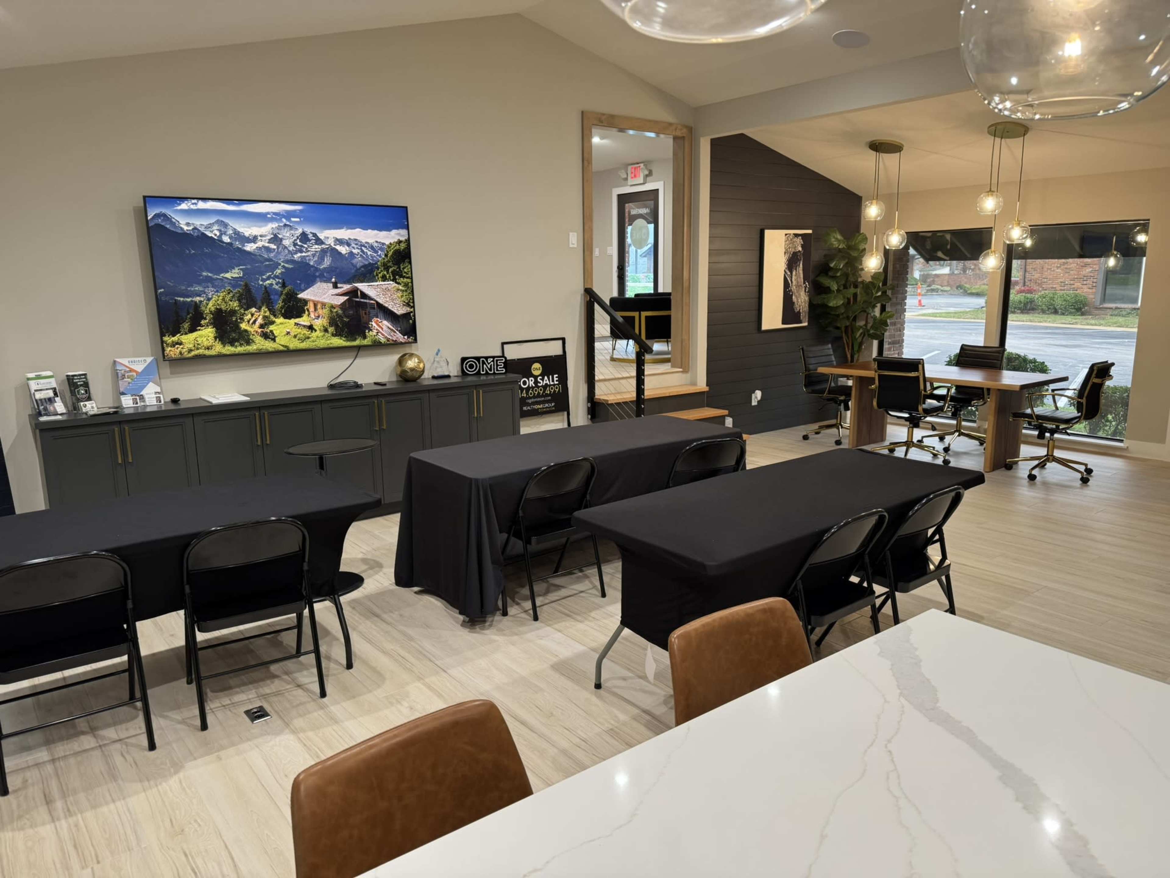 The image shows a modern conference room with several tables covered in black cloth, a television displaying a mountain landscape, and large windows letting in natural light.