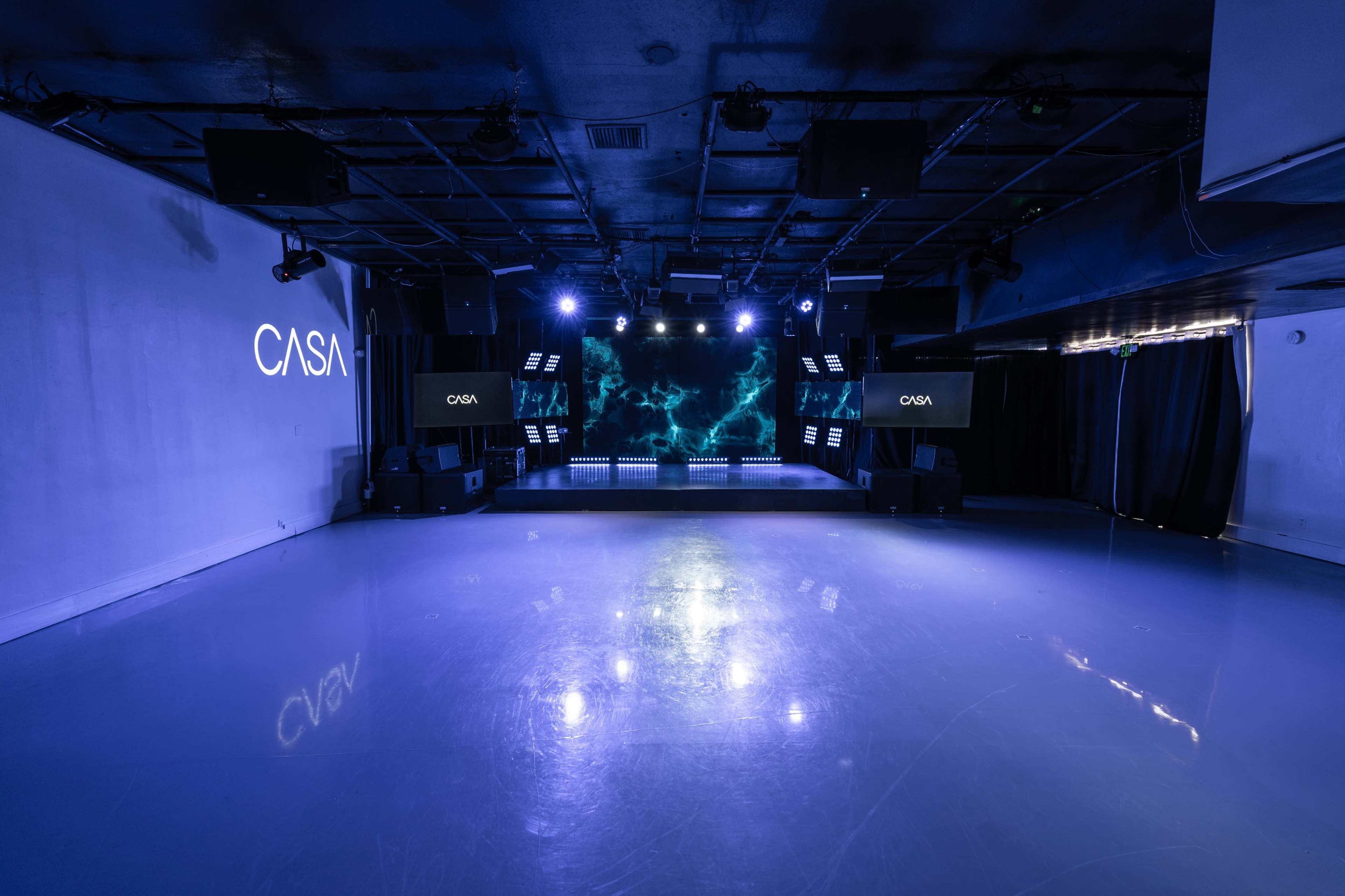 The image shows a modern event space with a shiny reflective floor, lit by blue lights and featuring a stage at the front.
