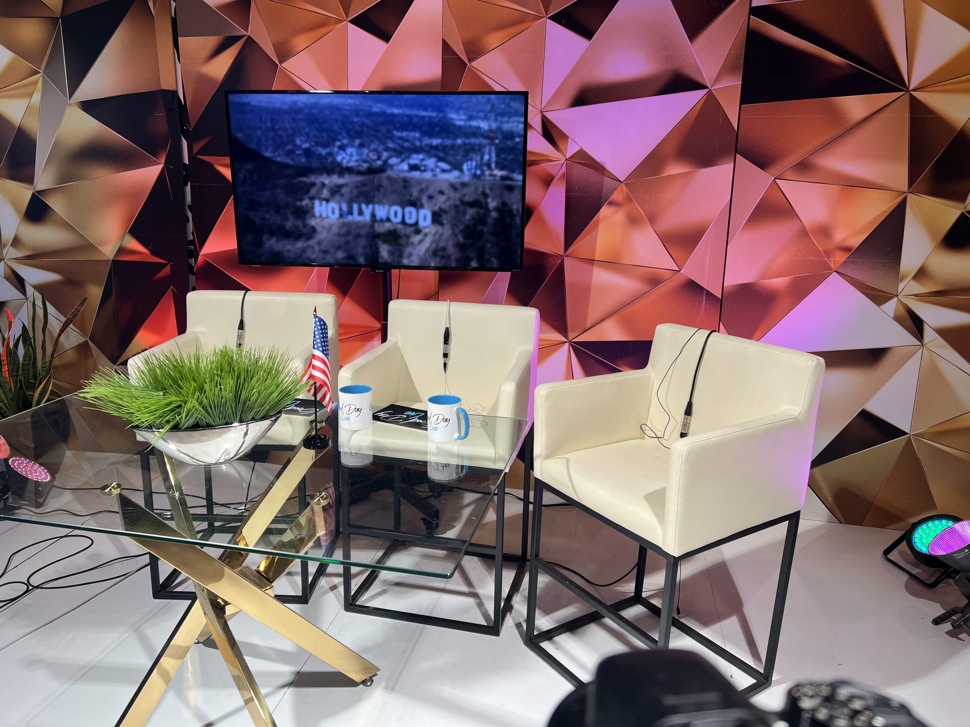 A television interview setup with three white chairs, a glass table, and a screen displaying "HOLLYWOOD" against a geometric backdrop.