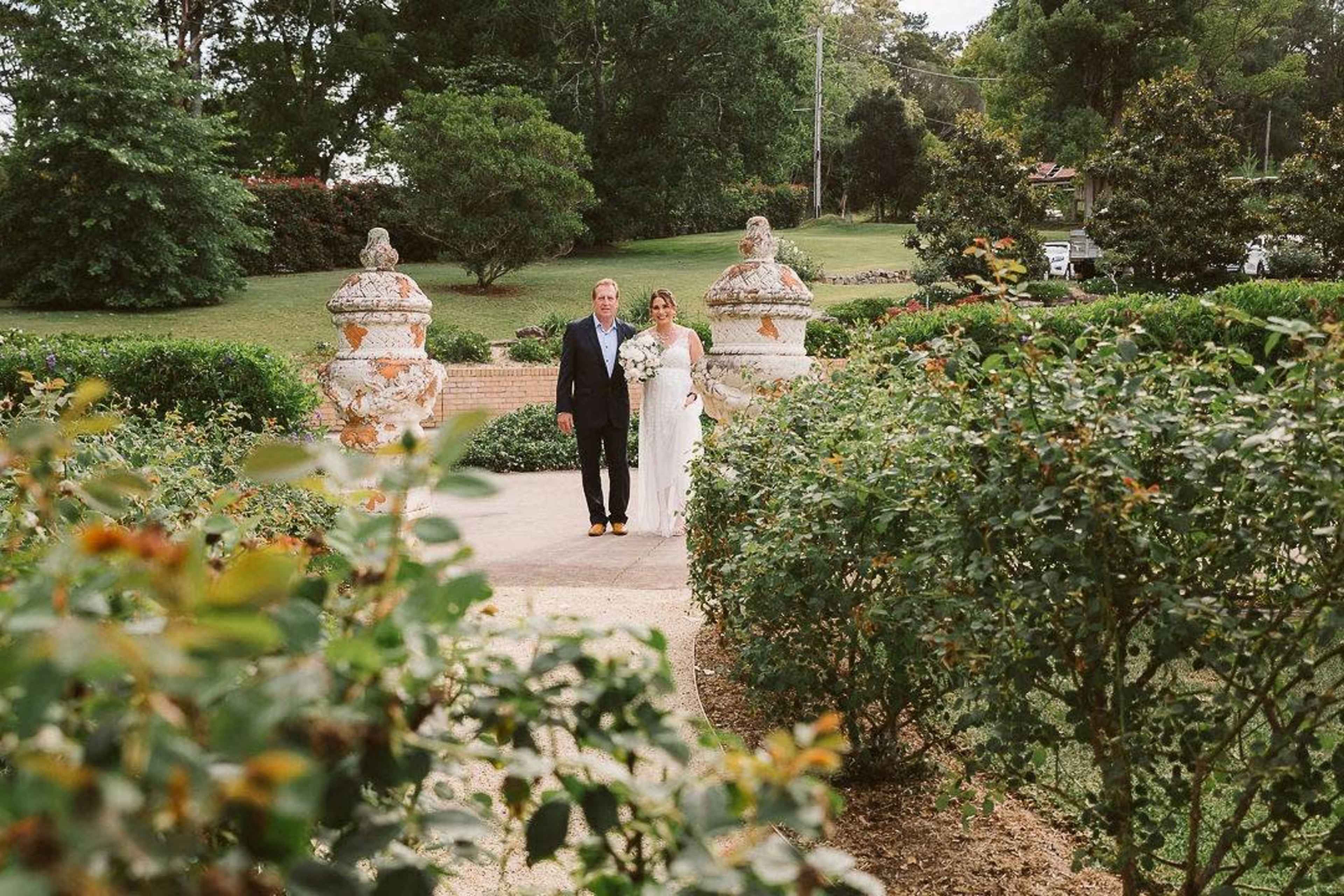 A couple stands together in a garden pathway surrounded by greenery and ornamental structures.