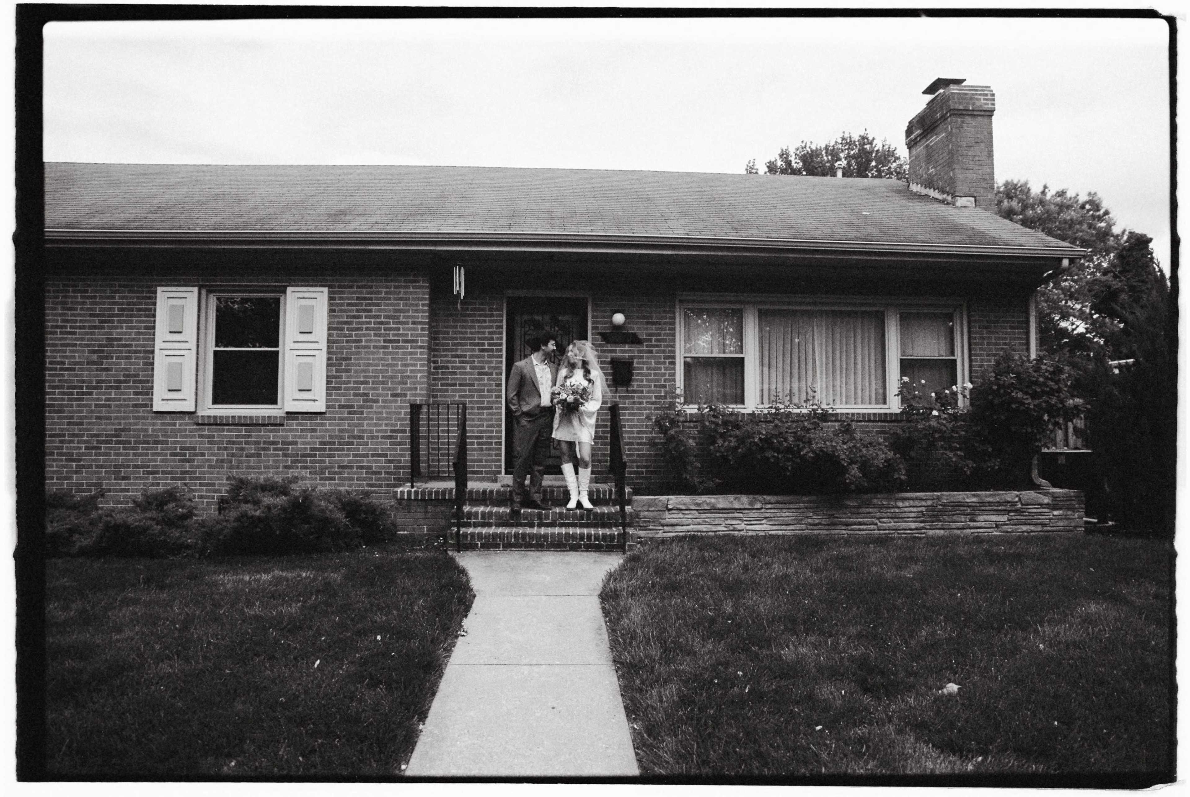 A couple stands together on the front steps of a brick house with a neatly manicured lawn.
