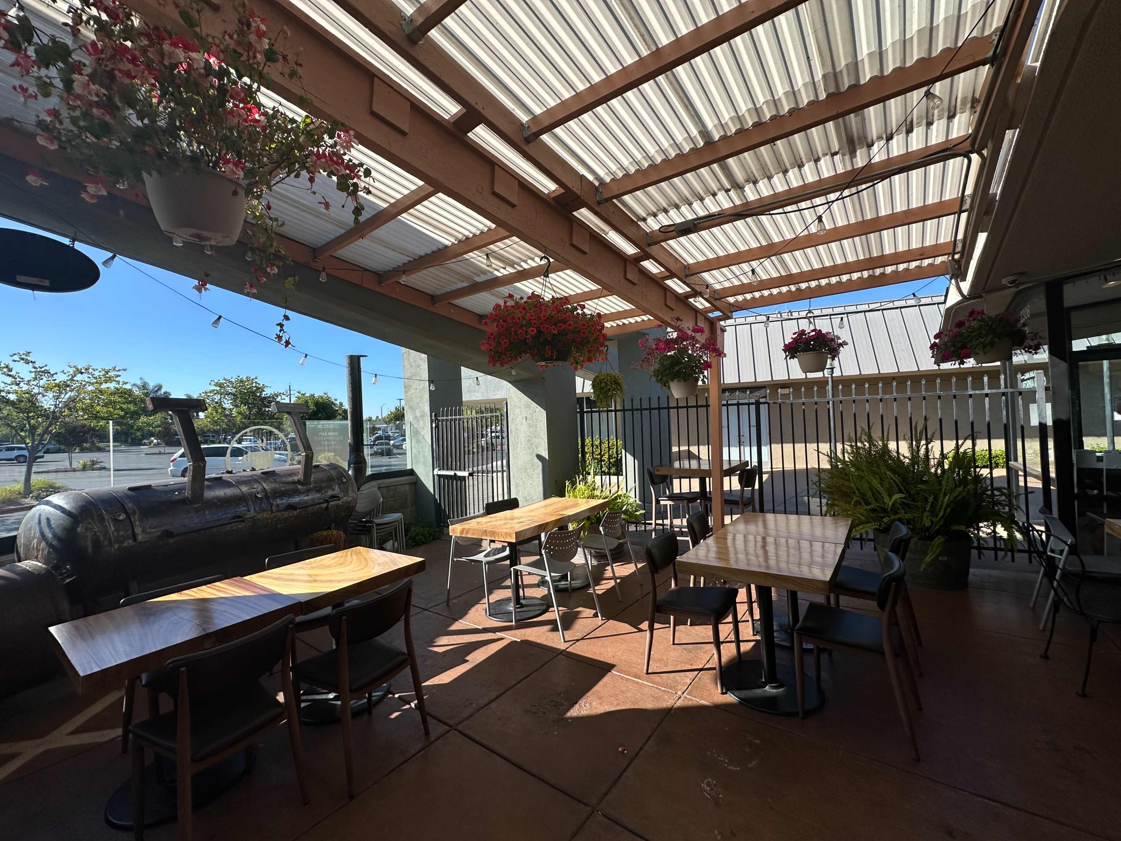 The image shows an outdoor dining area with wooden tables and chairs, covered by a pergola adorned with hanging flower pots.