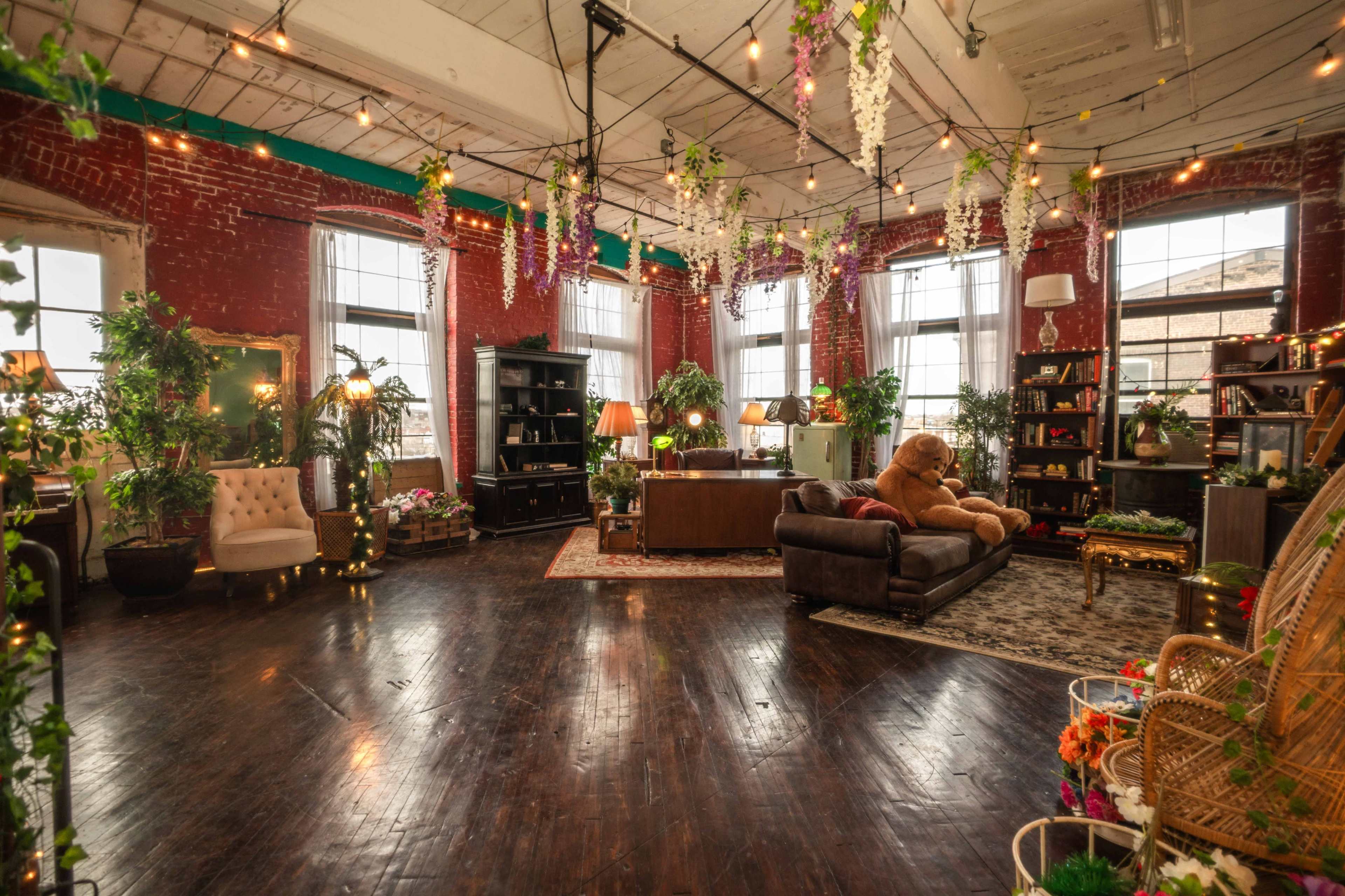 The image shows a spacious, decorated room with brick walls, wooden flooring, plants, and various furniture pieces, including a sofa and armchairs.
