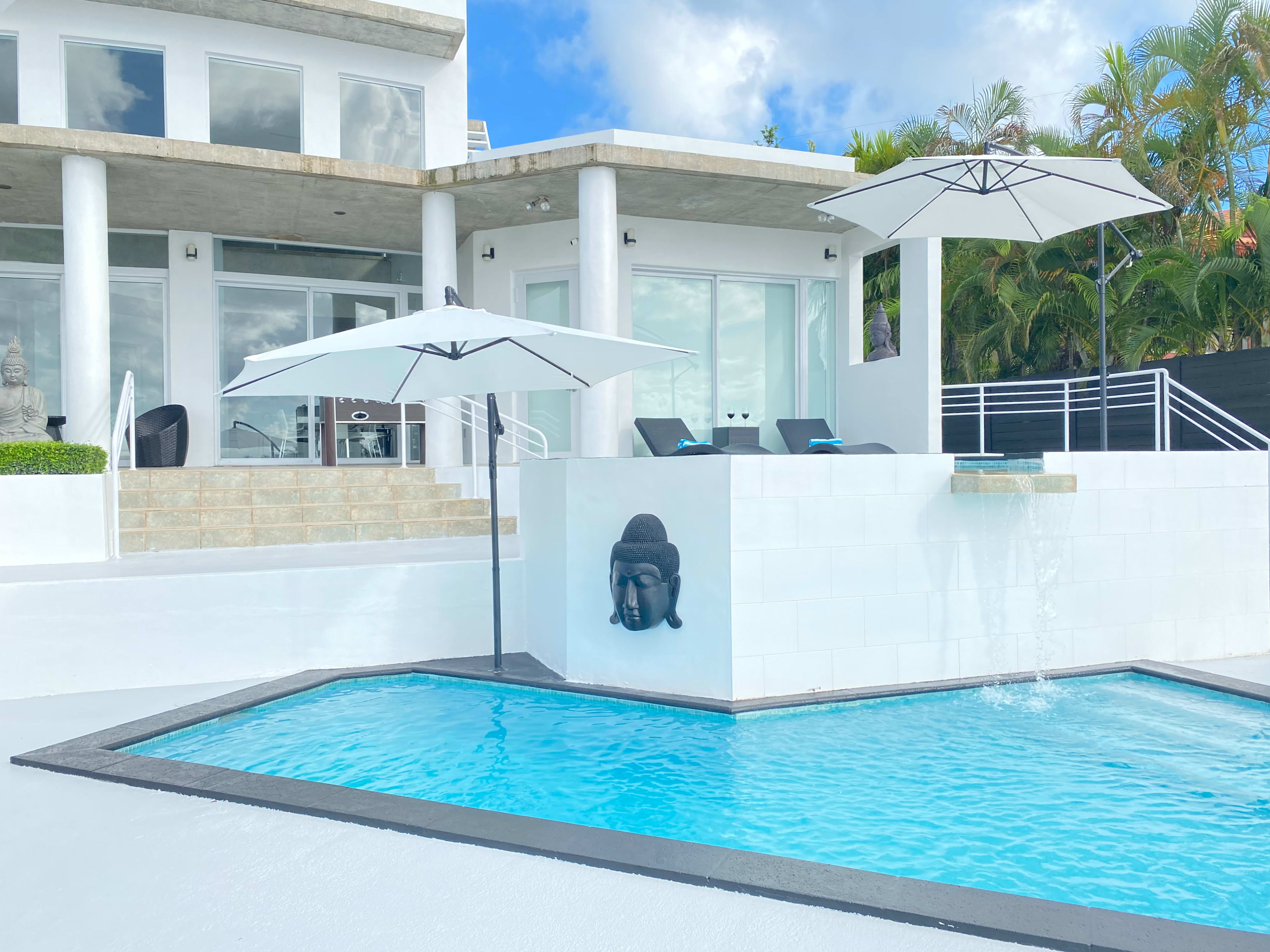 A modern pool area with a rectangular blue pool, white walls, a statue of Buddha, and two umbrellas positioned over lounge chairs outside a contemporary house.
