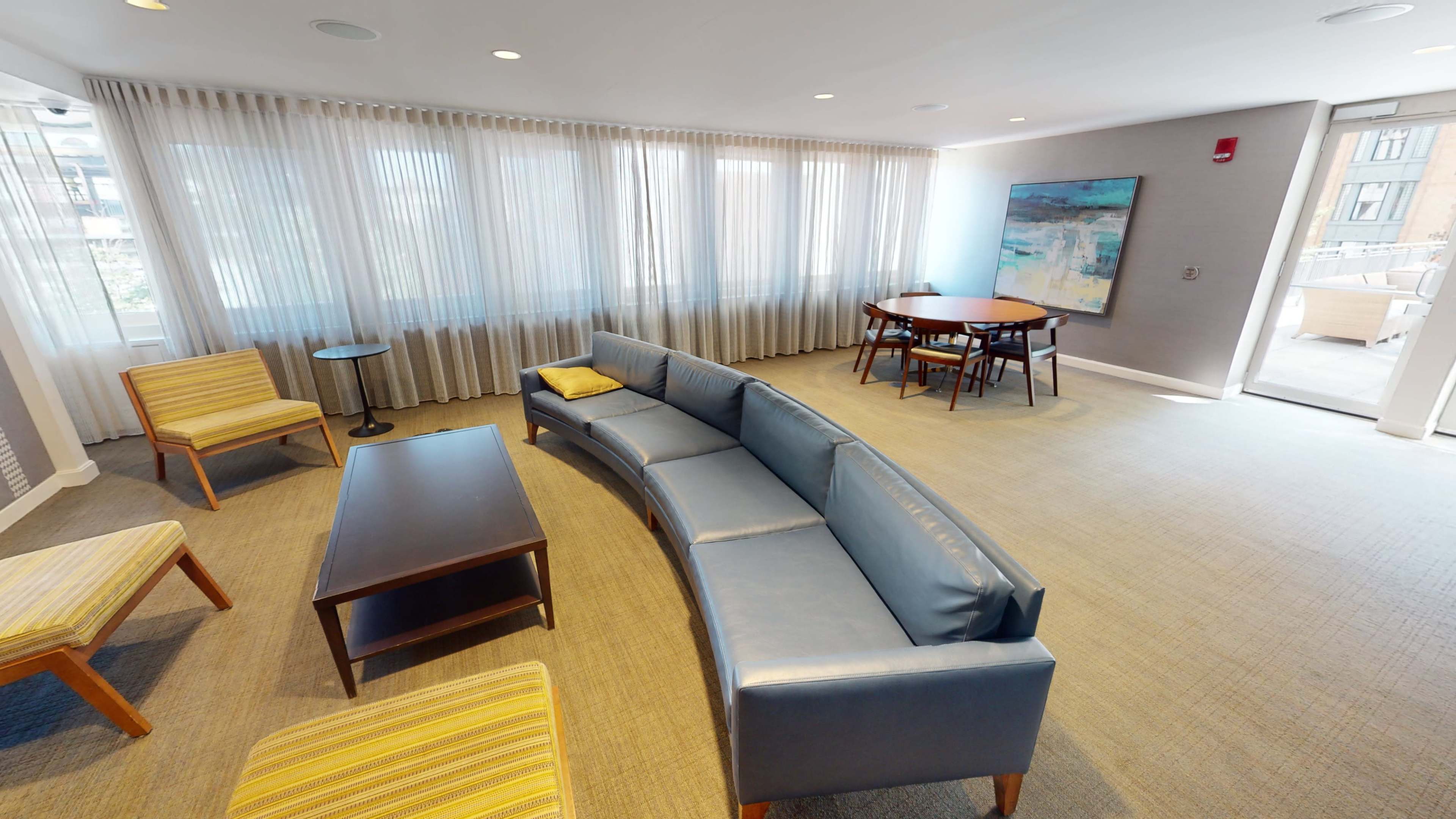 The scene features a spacious lounge area with a curved blue sofa, wooden chairs, a coffee table, and a dining table, all illuminated by natural light from large windows.