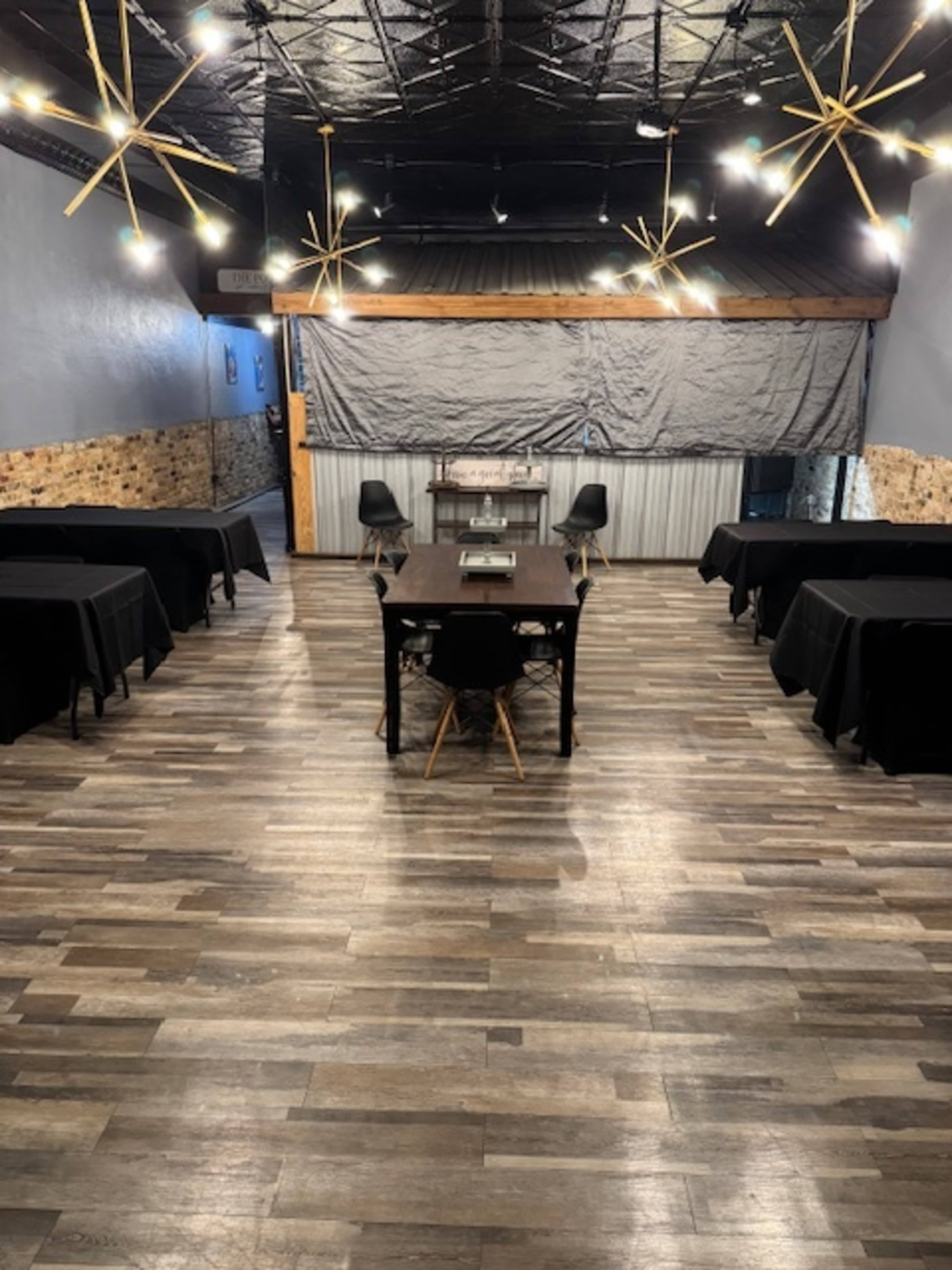 The image shows a spacious event room with several black-covered tables arranged evenly on a wooden floor, illuminated by overhead light fixtures.