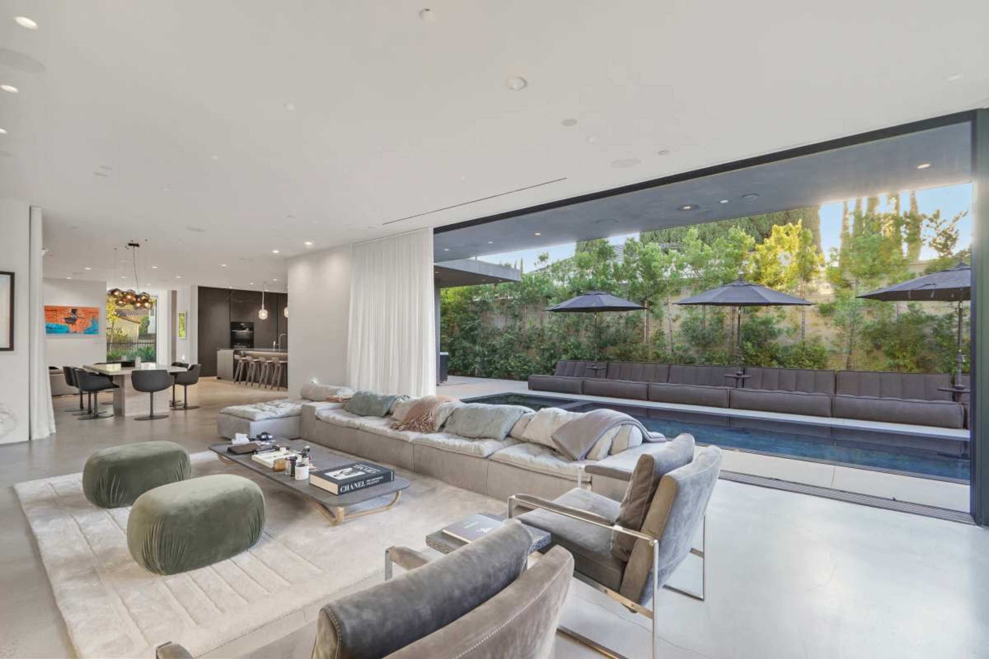 The image shows a modern living room with a large sectional sofa, plush seating, and an open view of an outdoor pool area surrounded by greenery.