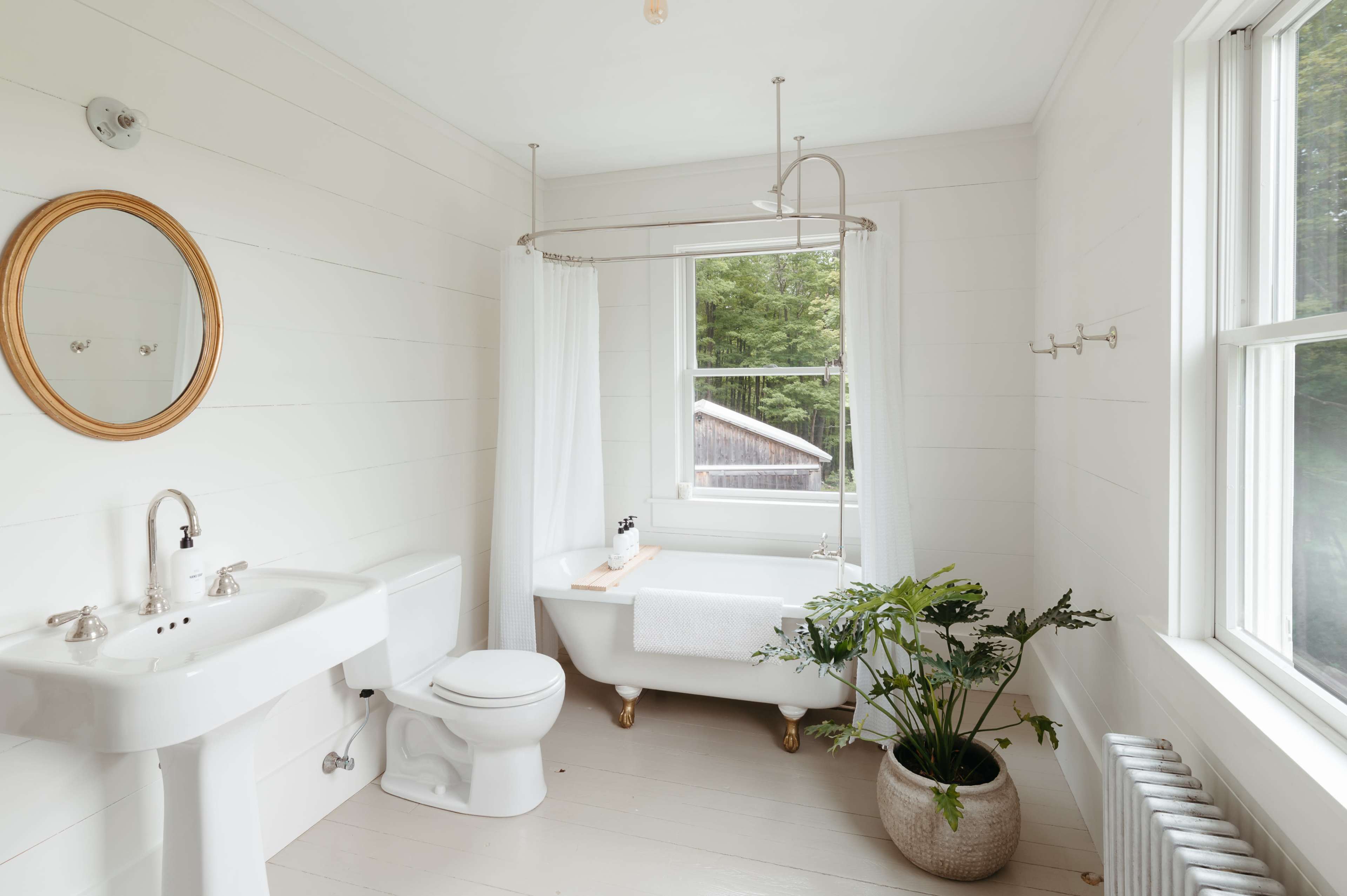 The bathroom features a freestanding bathtub, a pedestal sink, a toilet, and a large window with a view of greenery outside.