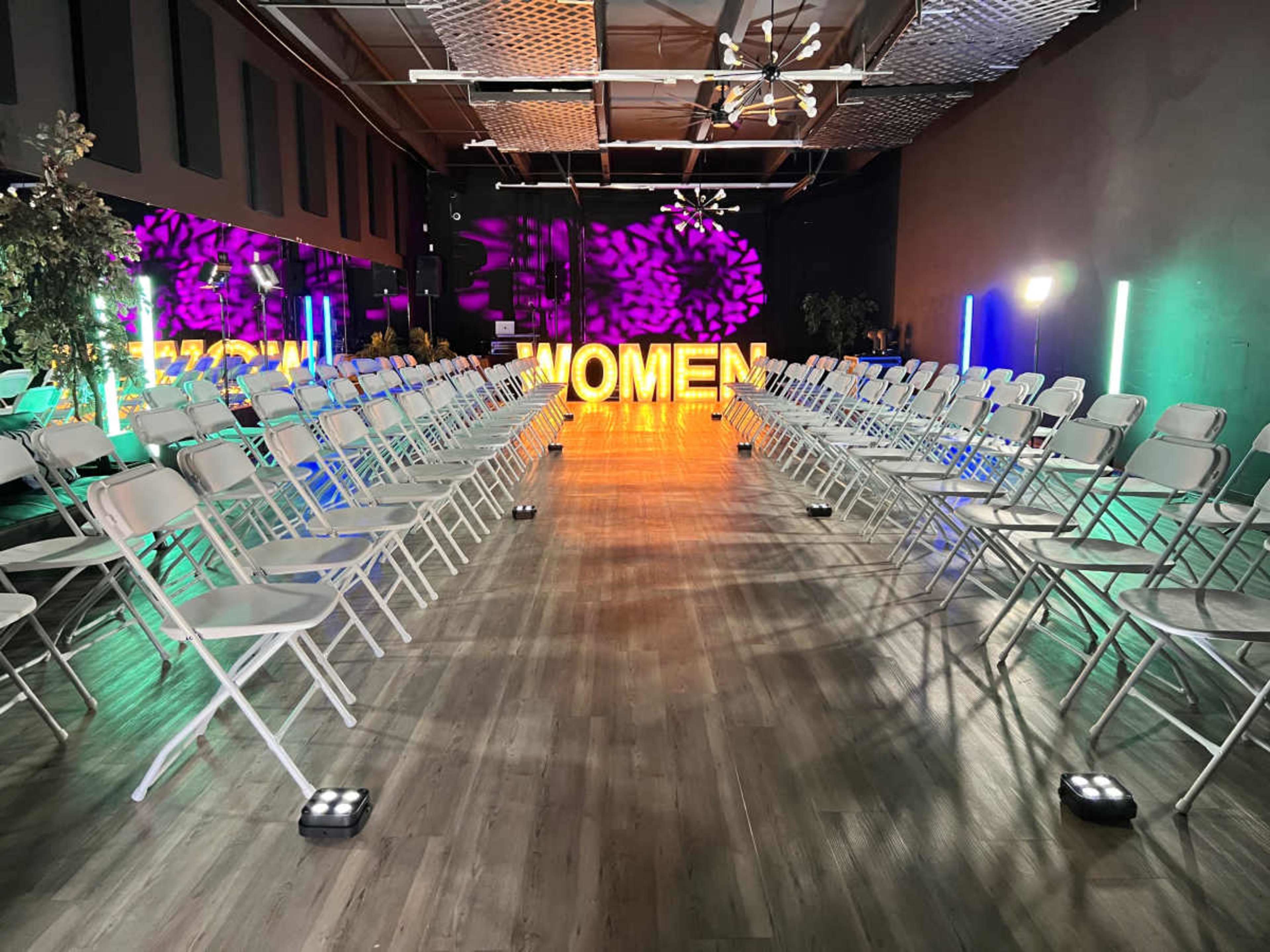 A spacious room is set up for an event, featuring rows of folding chairs facing a large illuminated sign that reads "WOMEN."